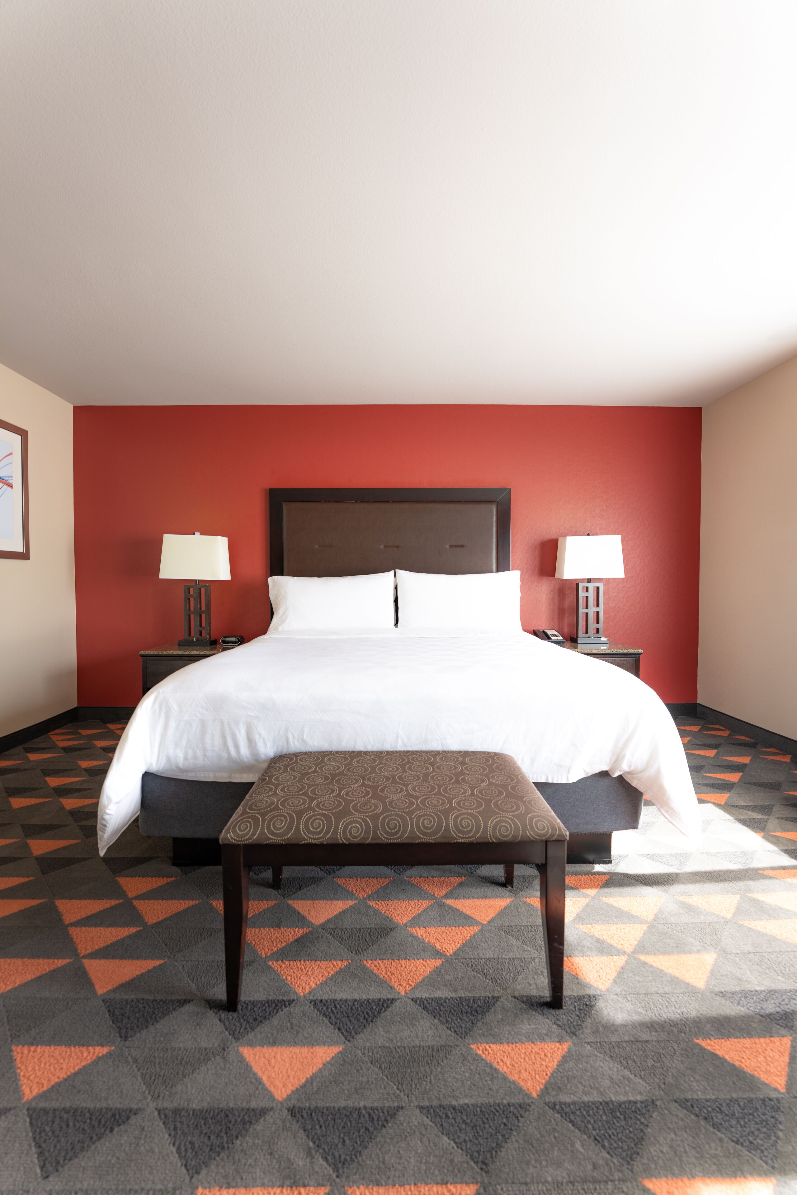 A hotel room with a large bed, two bedside tables with lamps, a patterned bench, and a geometric carpet.