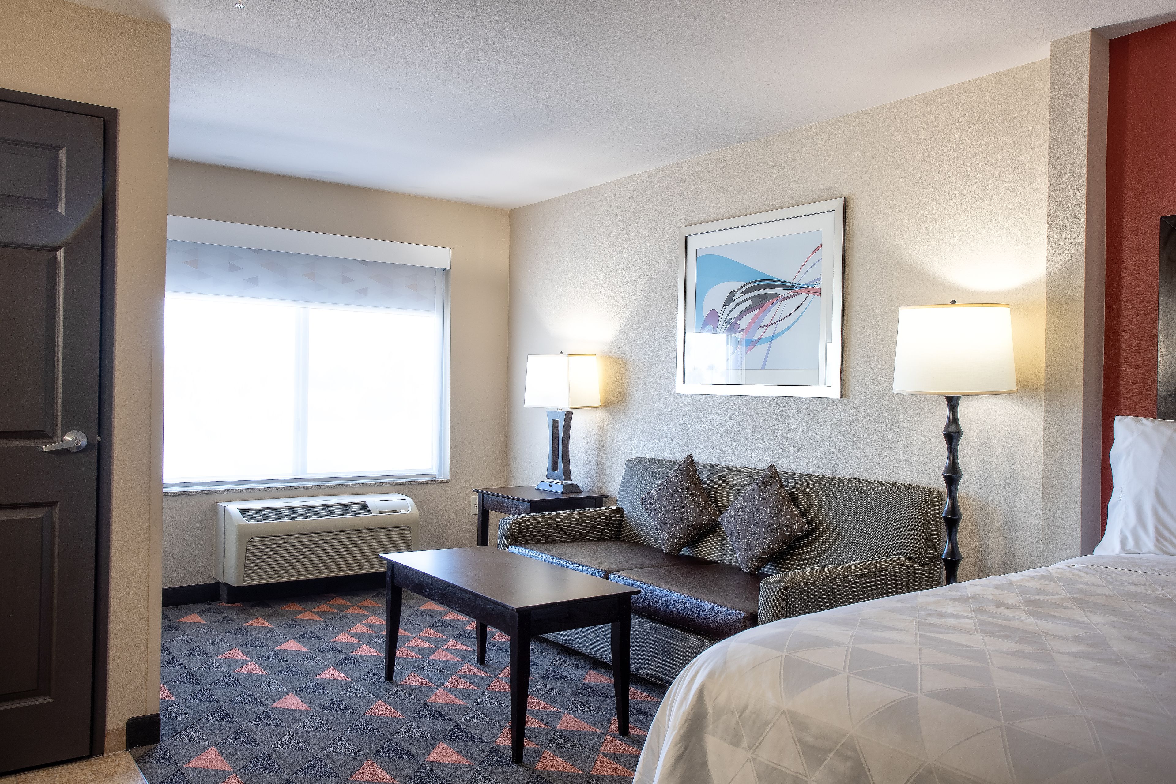 Habitación de hotel con cama, sofá, mesa, dos lámparas, una ventana con persianas y un radiador.