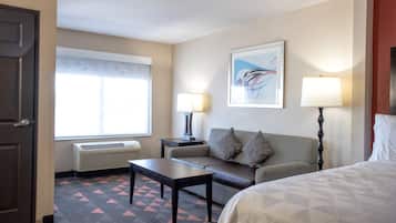 Habitación de hotel con cama, sofá, mesa, dos lámparas, una ventana con persianas y un radiador.