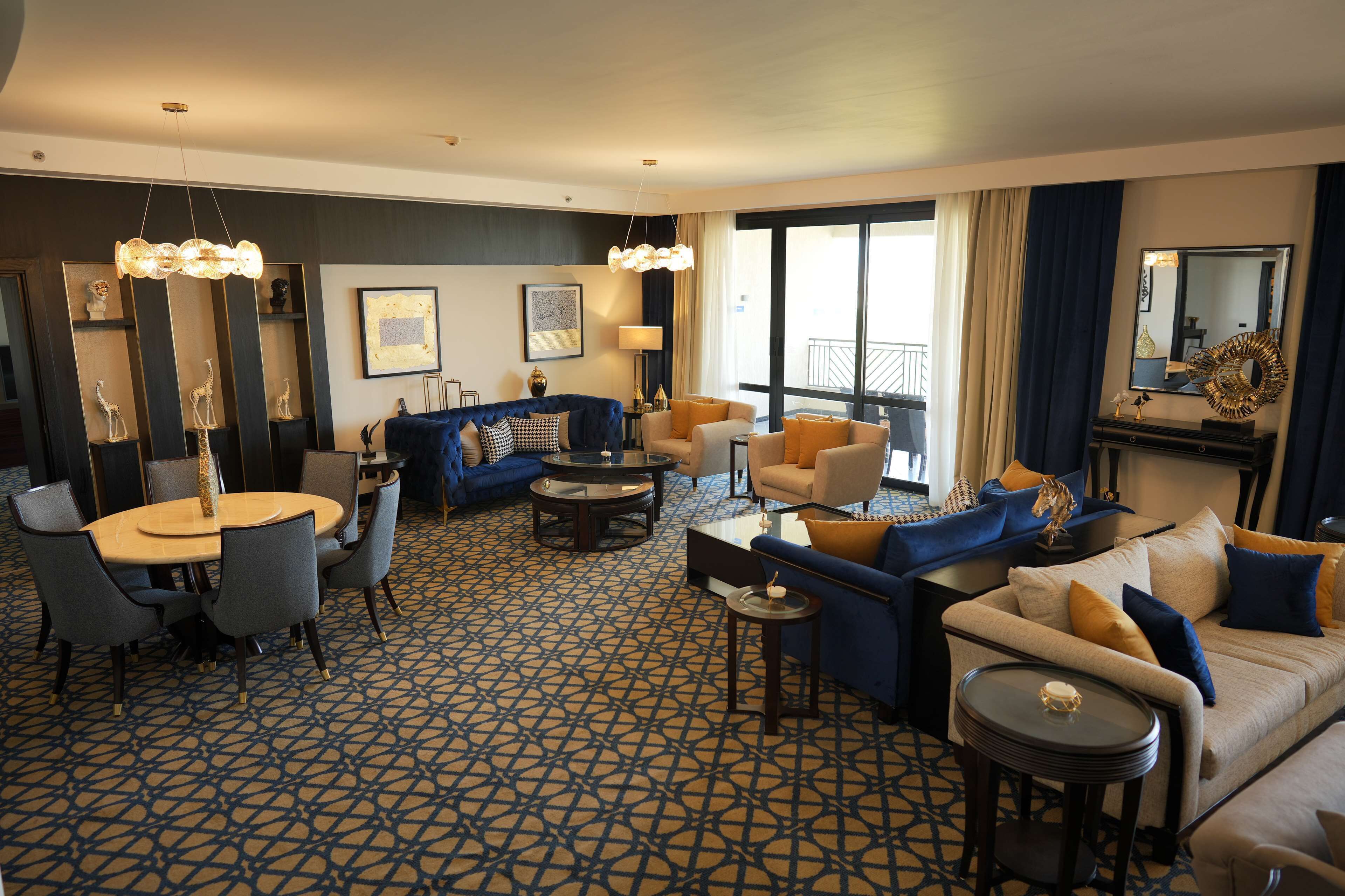 A modern hotel room with a round dining table, sofas, and a large window with curtains.