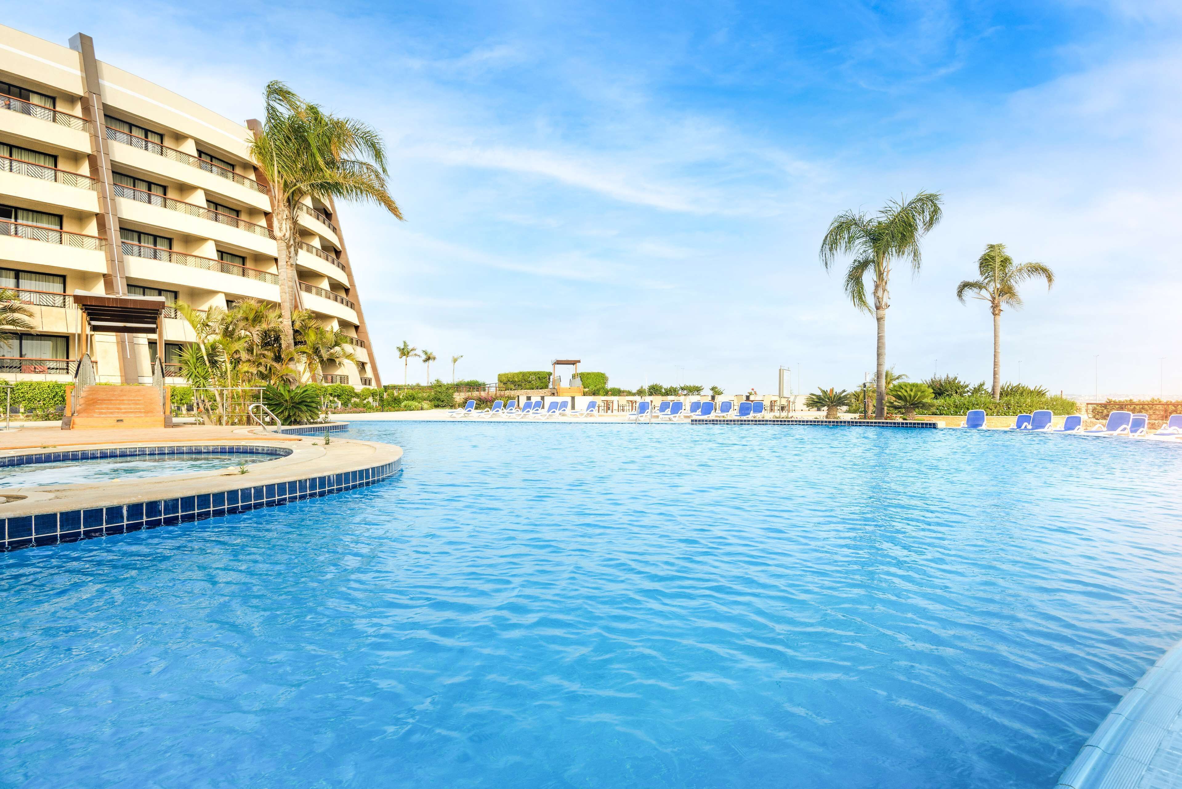 A swimming pool with lounge chairs and palm trees, surrounded by a hotel building.