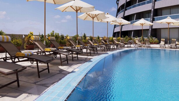 A pool area with lounge chairs and umbrellas, a tiled pool edge, and a modern building in the background. - Radisson Blu Hotel & Convention Center, Alexandria (Al-'Amriyah)