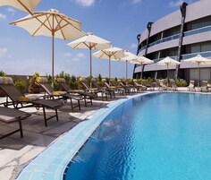 Un área de piscina con sillones de descanso y sombrillas, borde de piscina con azulejos y un edificio moderno al fondo.
