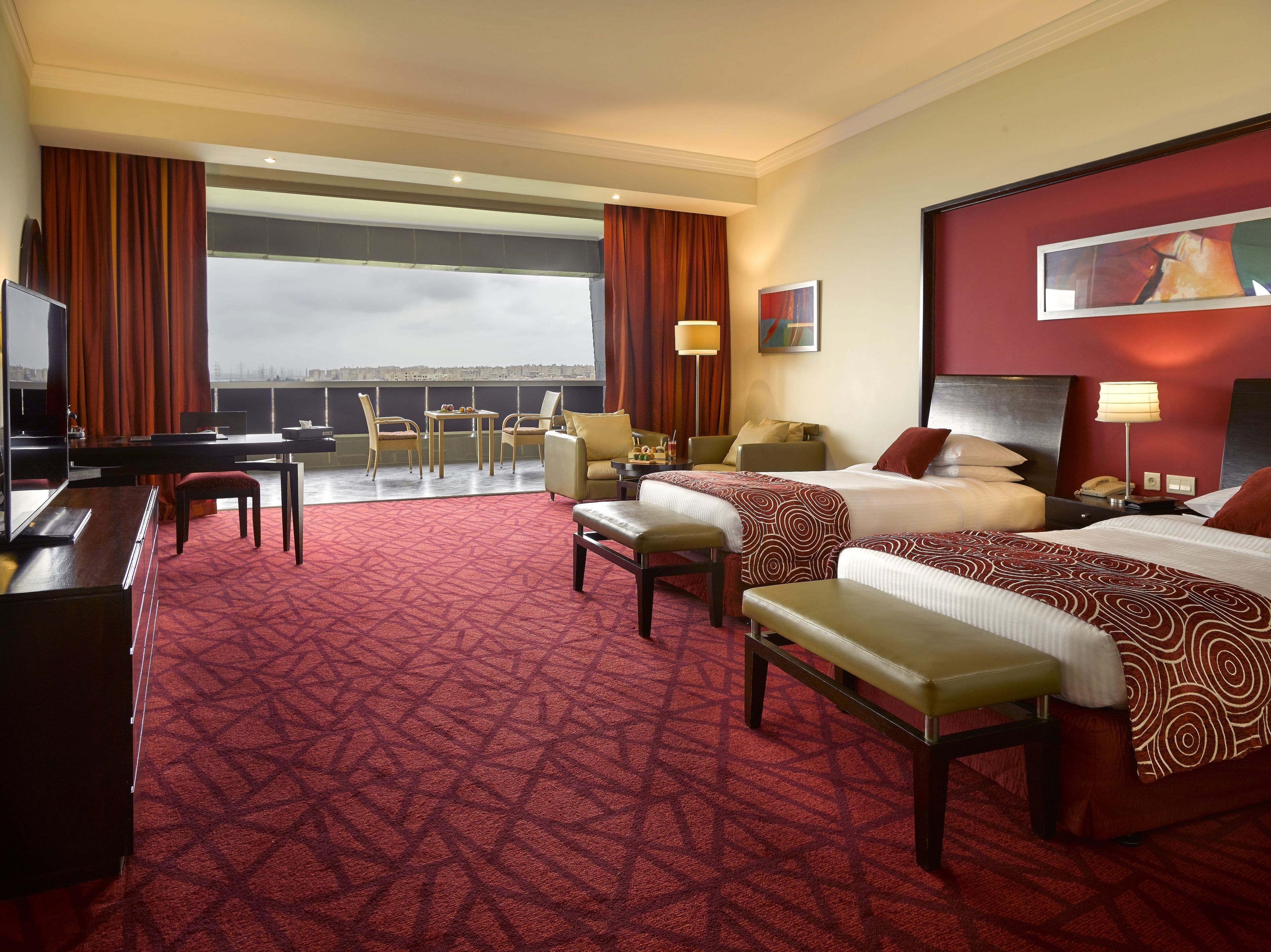 A hotel room with a large bed, two smaller beds, a desk, a chair, a TV, and a balcony with a city view.