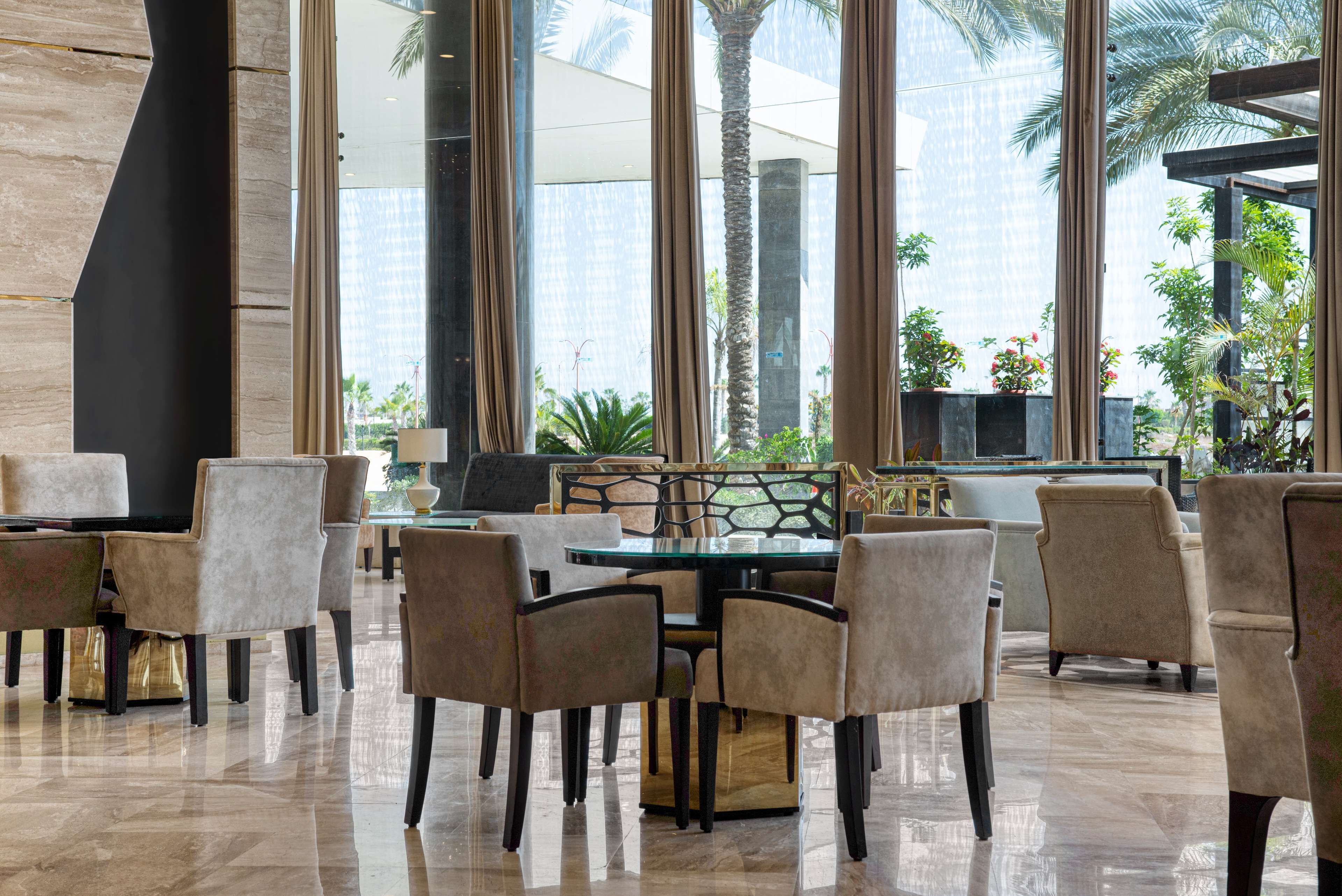 A modern hotel lobby with a glass coffee table surrounded by beige chairs, a stone pillar, and a view of palm trees and buildings.