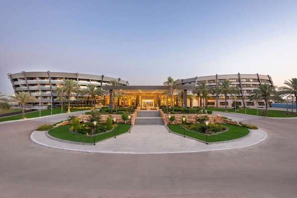 Un hotel moderno con fachada curva, entrada central con escalones y jardines ajardinados con palmeras.