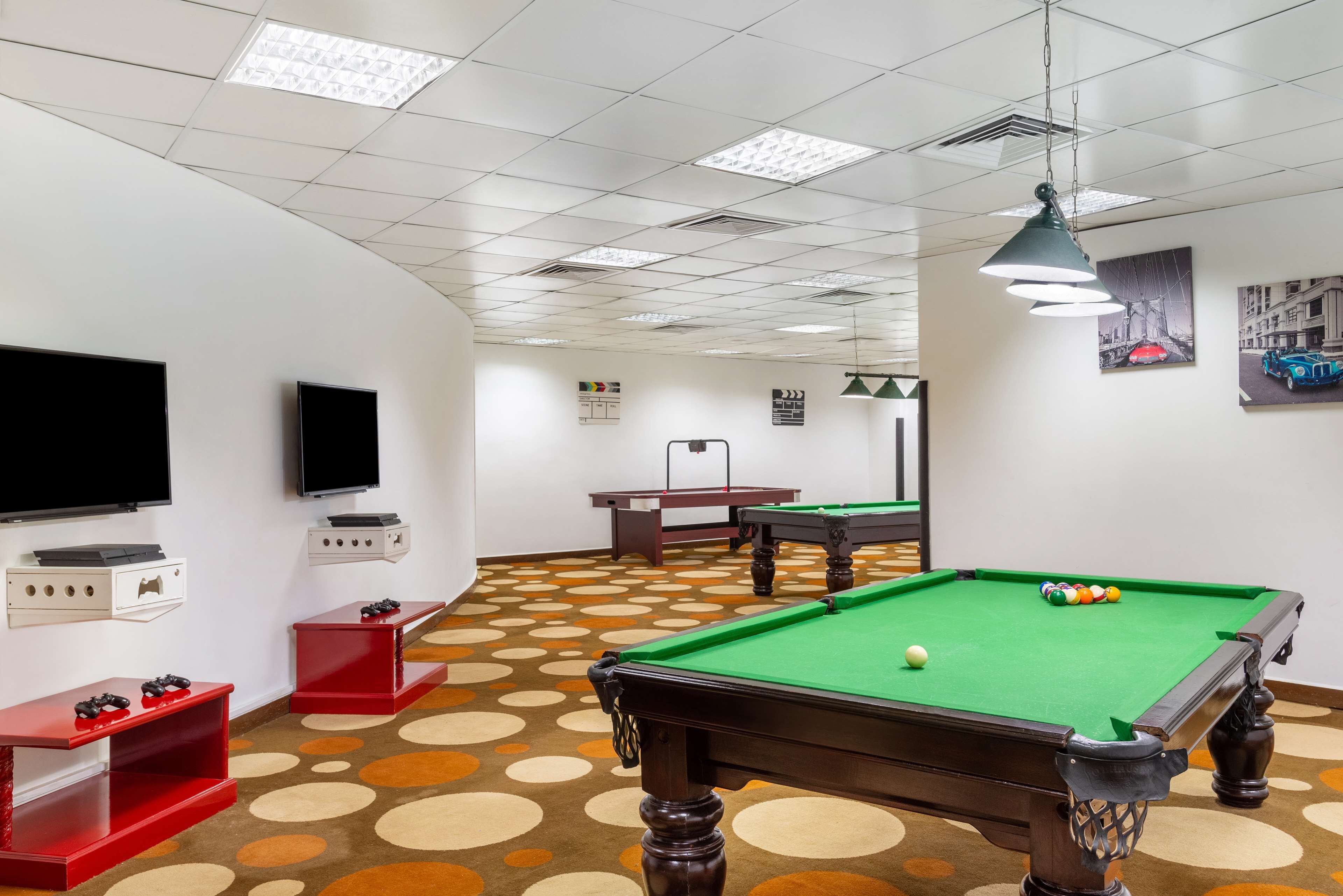 A pool table with a green felt surface, a pool cue, and a single white cue ball. There are two red entertainment units with game controllers, a television mounted on the wall, and a ceiling with fluorescent lighting.