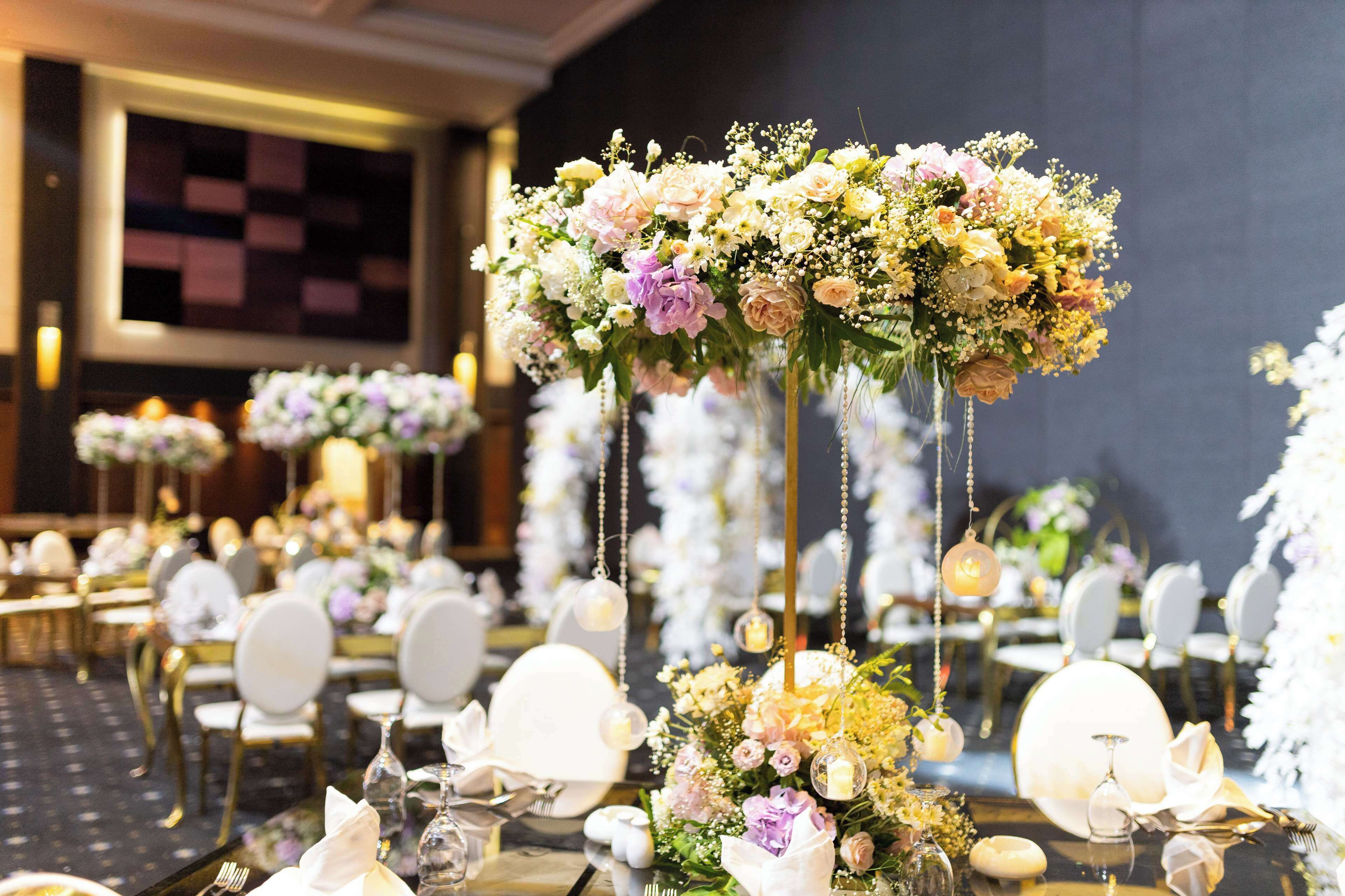 A banquet hall with round tables set for a formal event, featuring floral centerpieces and hanging decorations.