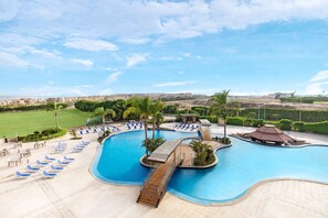 Piscina de un resort con un puente de madera, sillas de descanso y vista a la ciudad de fondo.