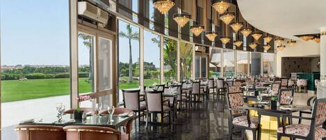 Un comedor espacioso con varias mesas y sillas, ventanas amplias que ofrecen vistas al exterior, y elegantes candelabros colgados del techo.