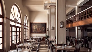 Ein geräumiges Restaurant mit hohen Decken, großen Fenstern und einem zentral platzierten Gemälde. Die Tische sind ordentlich mit Stühlen angerichtet, und es gibt eine Empore mit zusätzlichen Sitzmöglichkeiten.