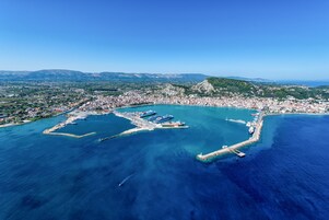 Vista aerea di una città costiera con un porto turistico, acque blu cristalline e montagne sullo sfondo.