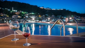 Ein Tisch am Pool mit zwei Weingläsern, Blick auf ein Schwimmbad und eine Stadt am Hang in der Abenddämmerung.