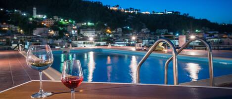 Mesa à beira da piscina com duas taças de vinho, com vista para uma piscina e uma cidade situada numa colina ao anoitecer.