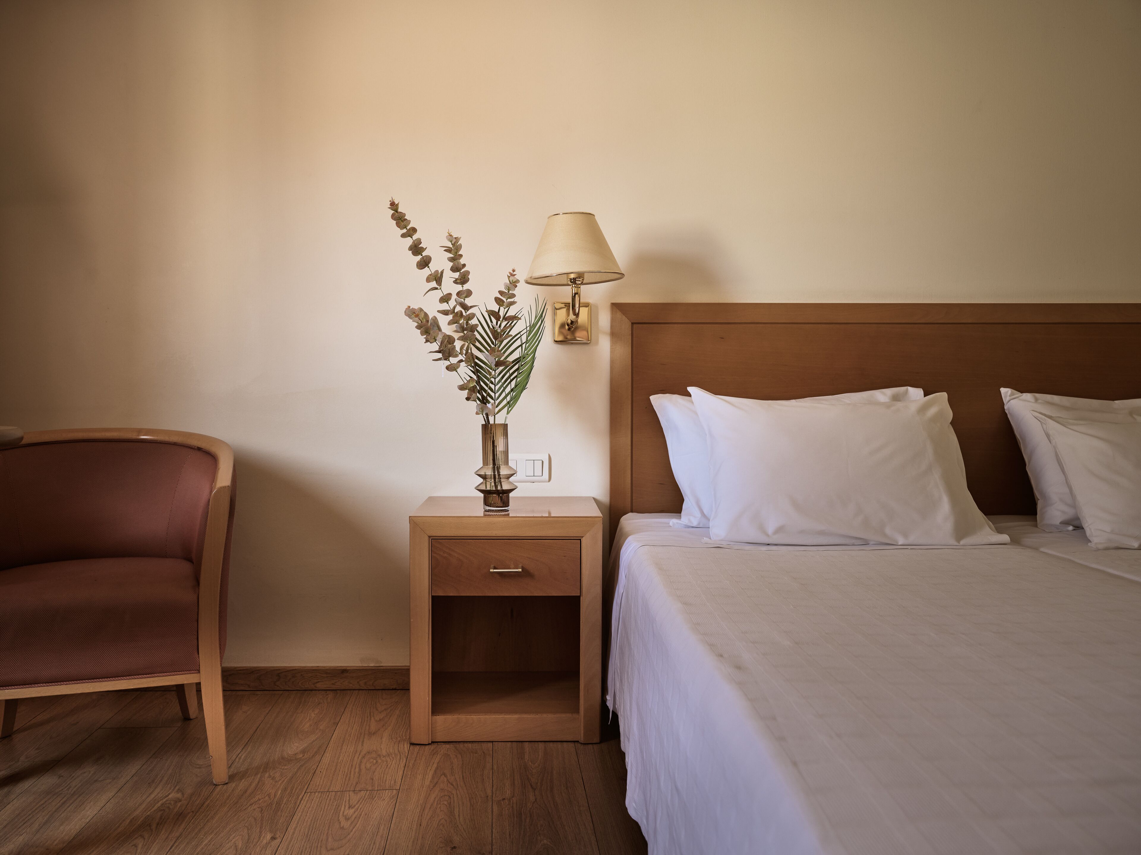 Une chambre d’hôtel avec une table de chevet en bois, une lampe, un vase contenant des plantes séchées, un lit avec du linge de lit blanc et une chaise en bois.