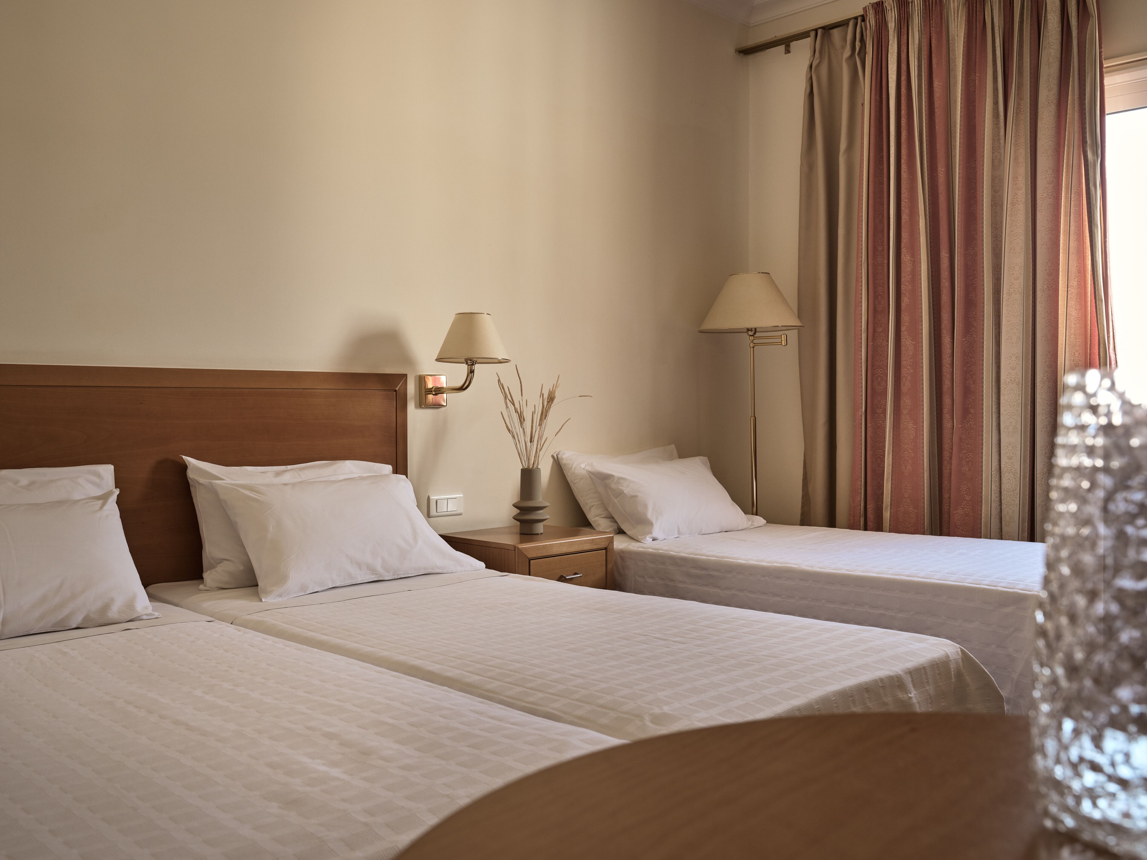 A hotel room with two single beds, a nightstand with a lamp, and a window with curtains.