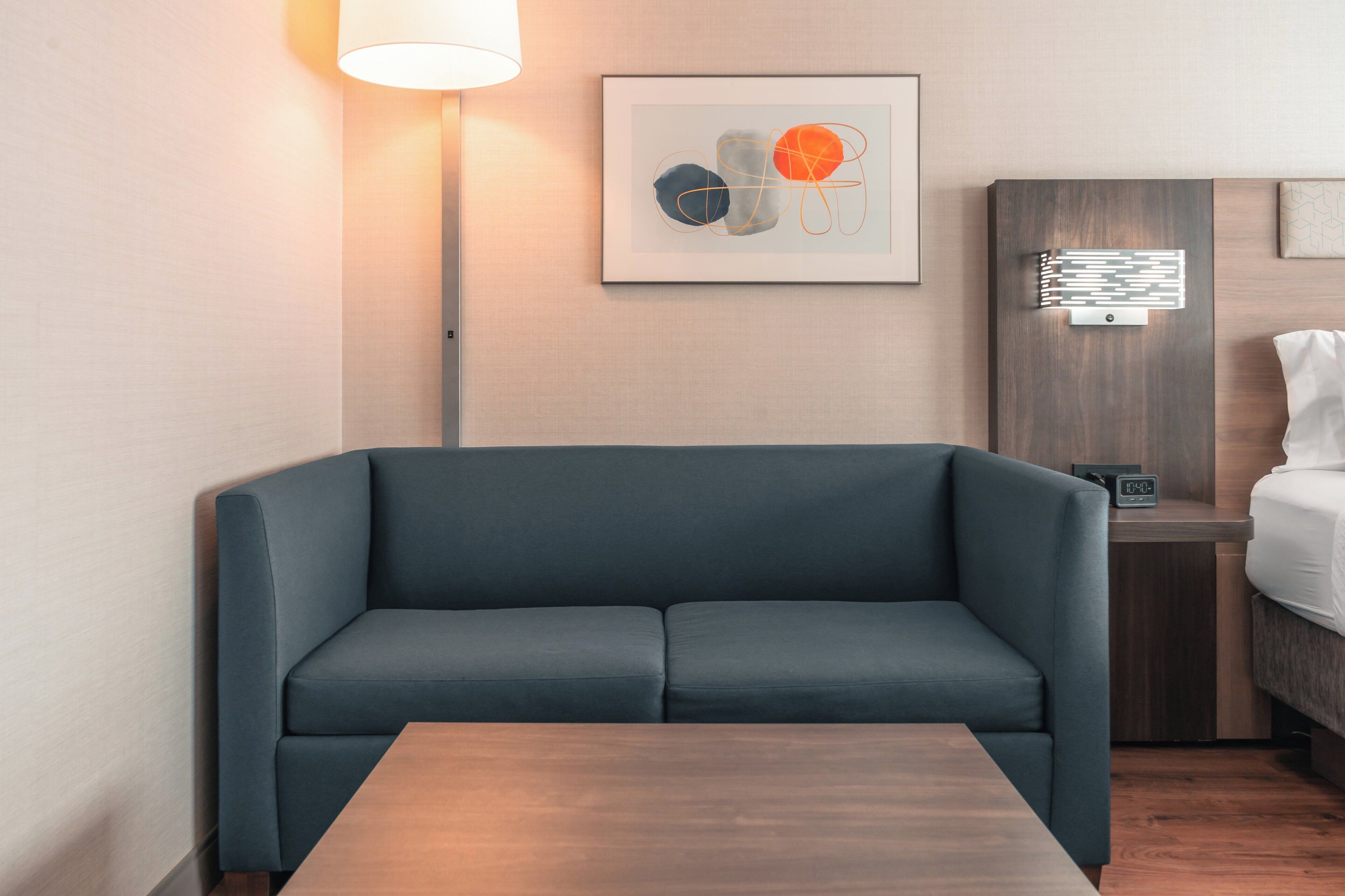 A modern room with a dark blue sofa, a wooden table, a bedside table with a lamp, and a framed abstract art piece on the wall.