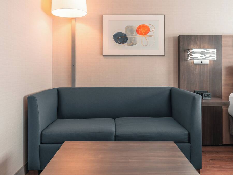 A modern room with a dark blue sofa, a wooden table, a bedside table with a lamp, and a framed abstract art piece on the wall.