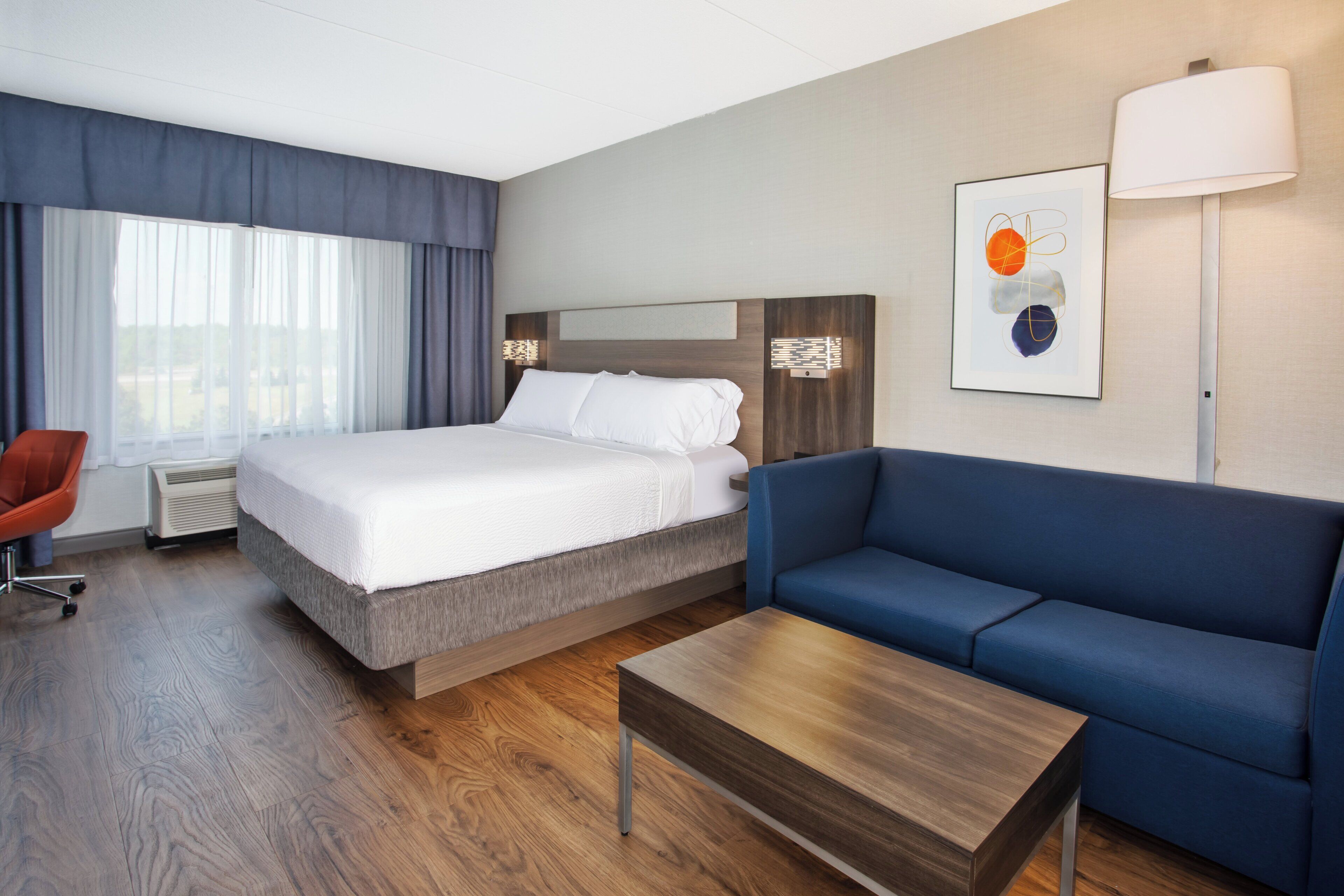 A hotel room with a bed, a blue sofa, a wooden coffee table, a red chair, and a large window with curtains.