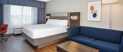 A hotel room with a bed, a blue sofa, a wooden coffee table, a red chair, and a large window with curtains.