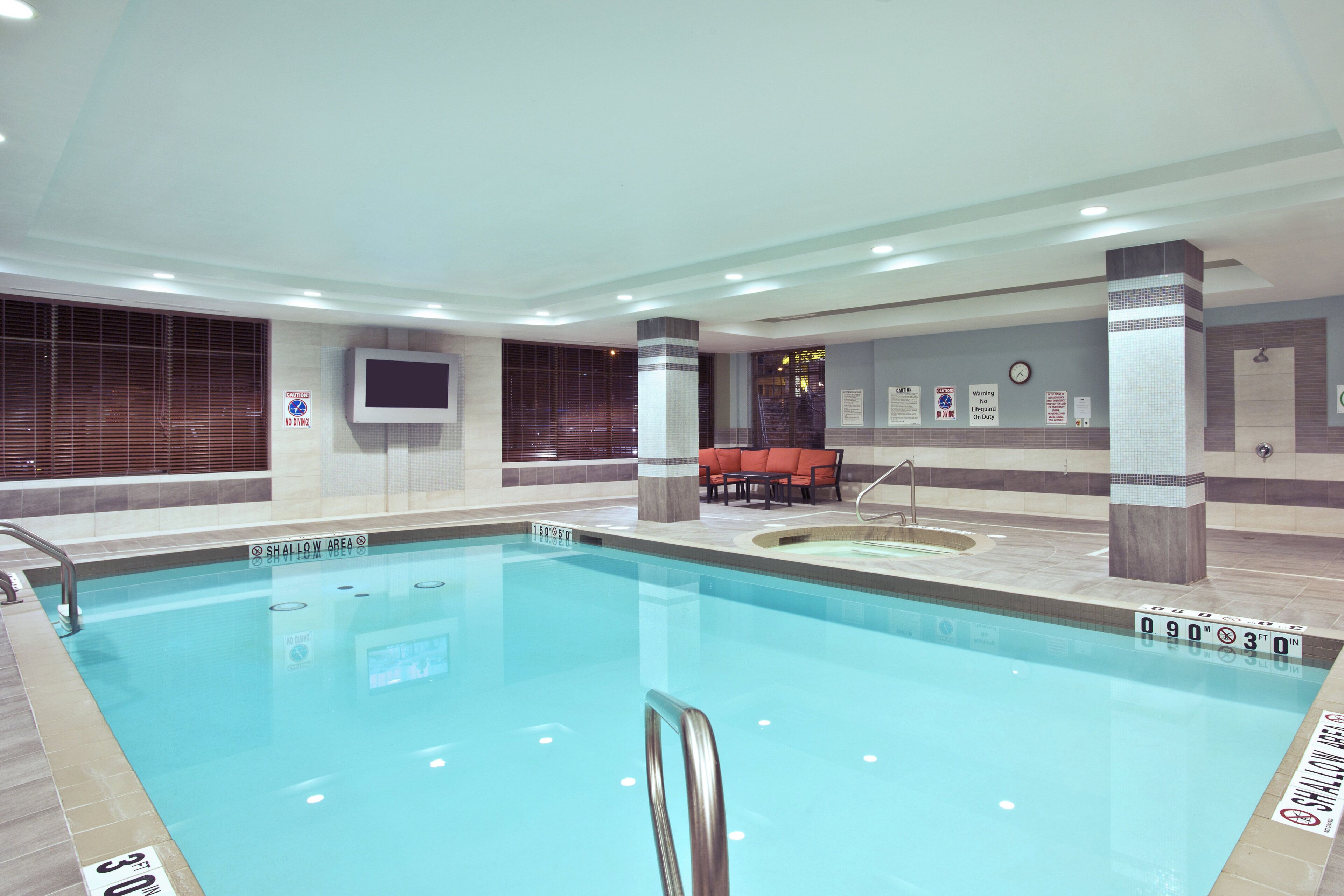 An indoor swimming pool with a shallow area marked, a hot tub, and seating area with tables and chairs.