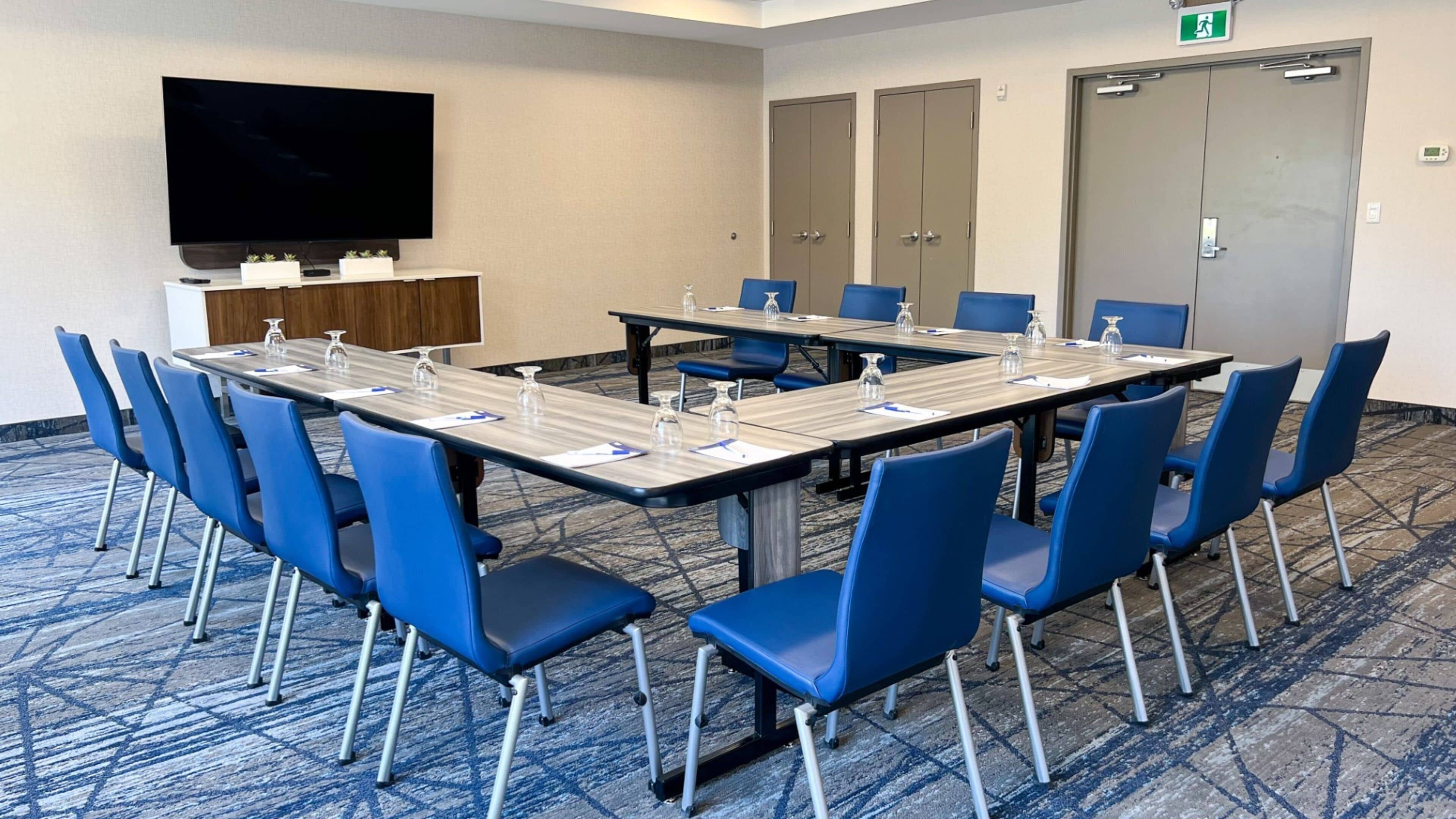 A conference room with a long table, blue chairs, a flat-screen TV, a small kitchenette, and a carpeted floor.