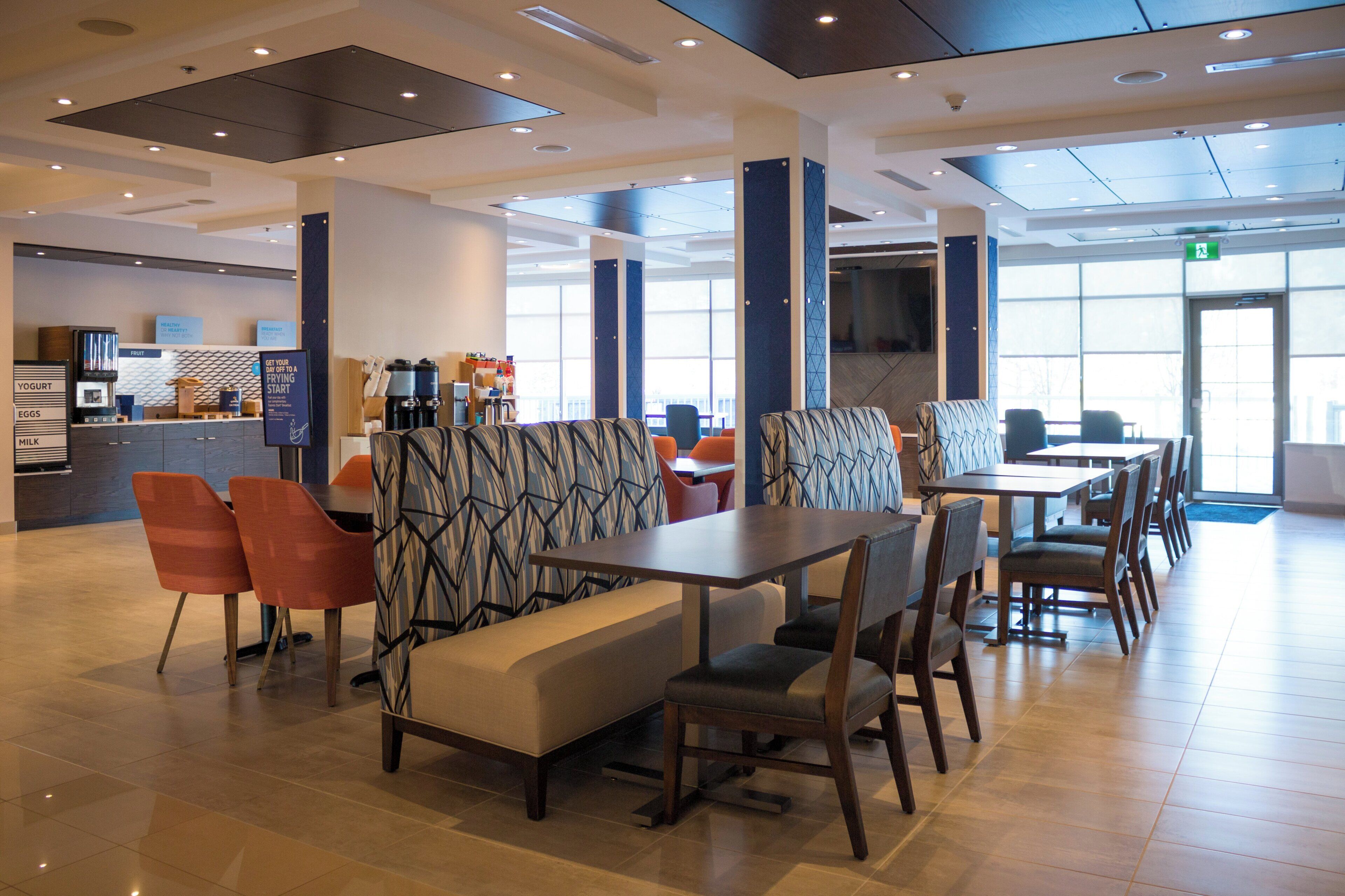 A modern dining area with high tables and chairs, a counter with breakfast options, and large windows allowing natural light.