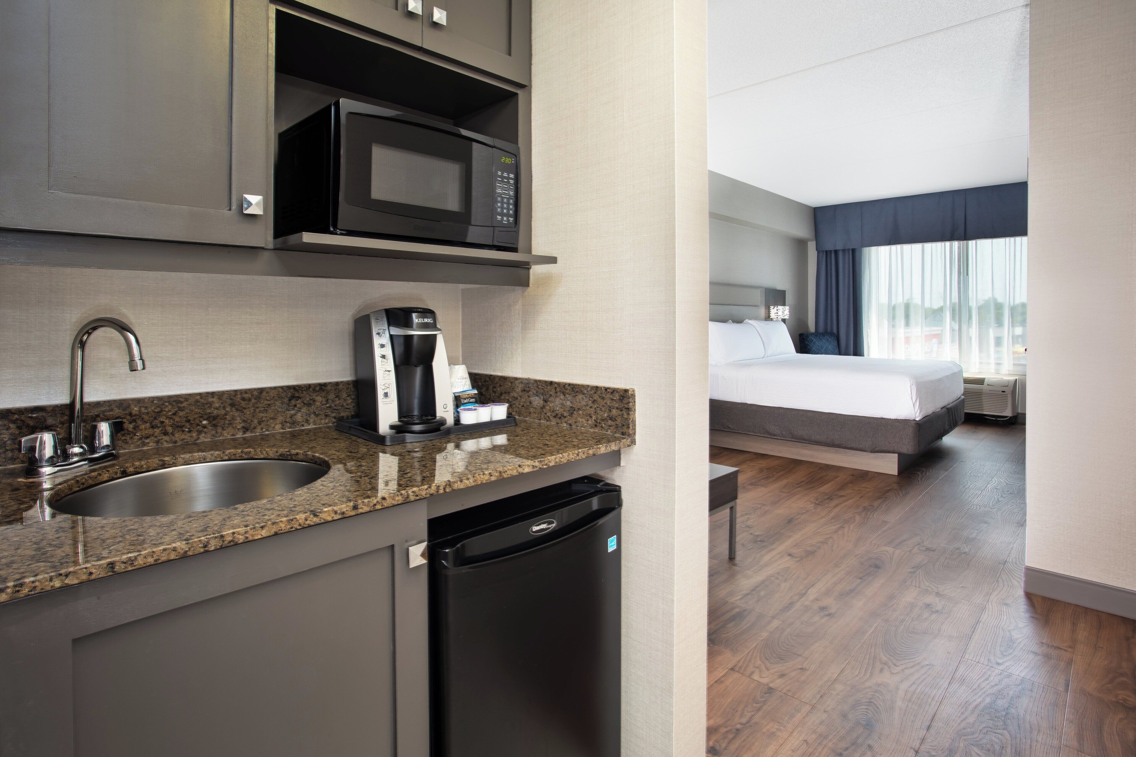 A modern hotel room with a kitchen area including a microwave, coffee maker, sink, and dishwasher.