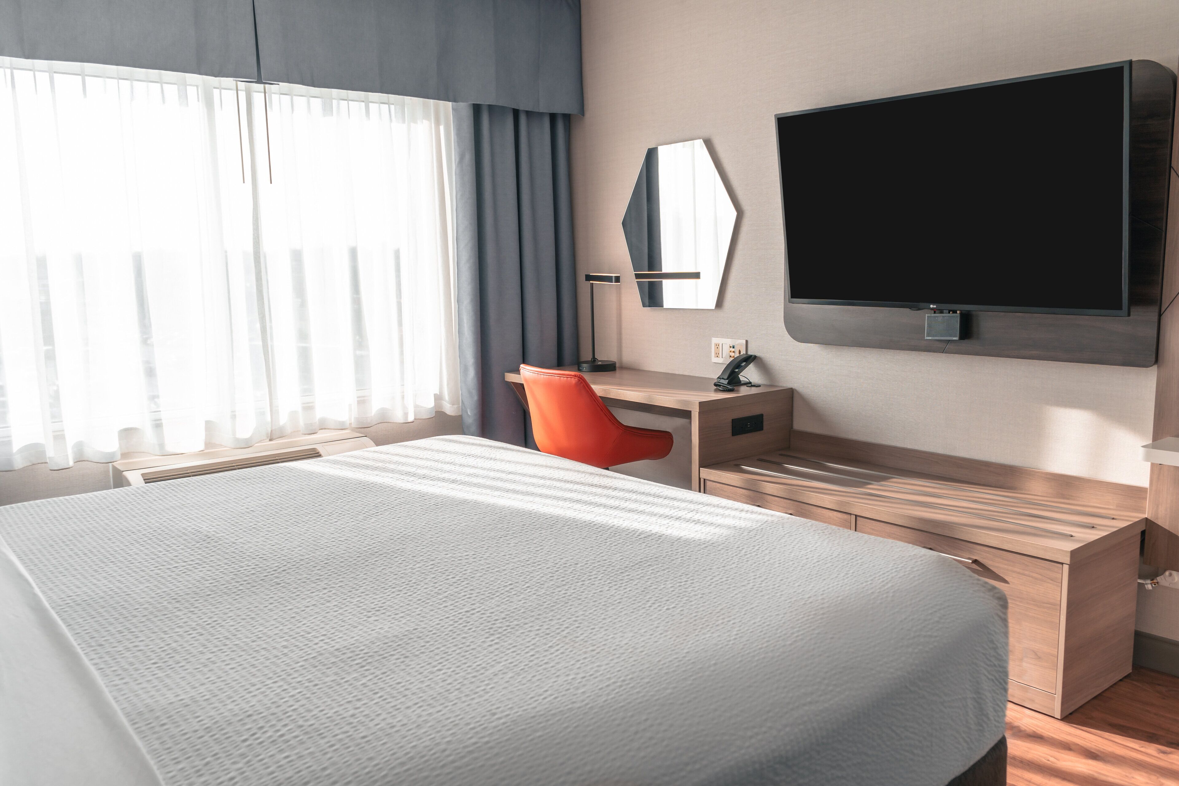 A hotel room with a large bed, a desk with a chair, a flat-screen TV, and a large window with curtains.