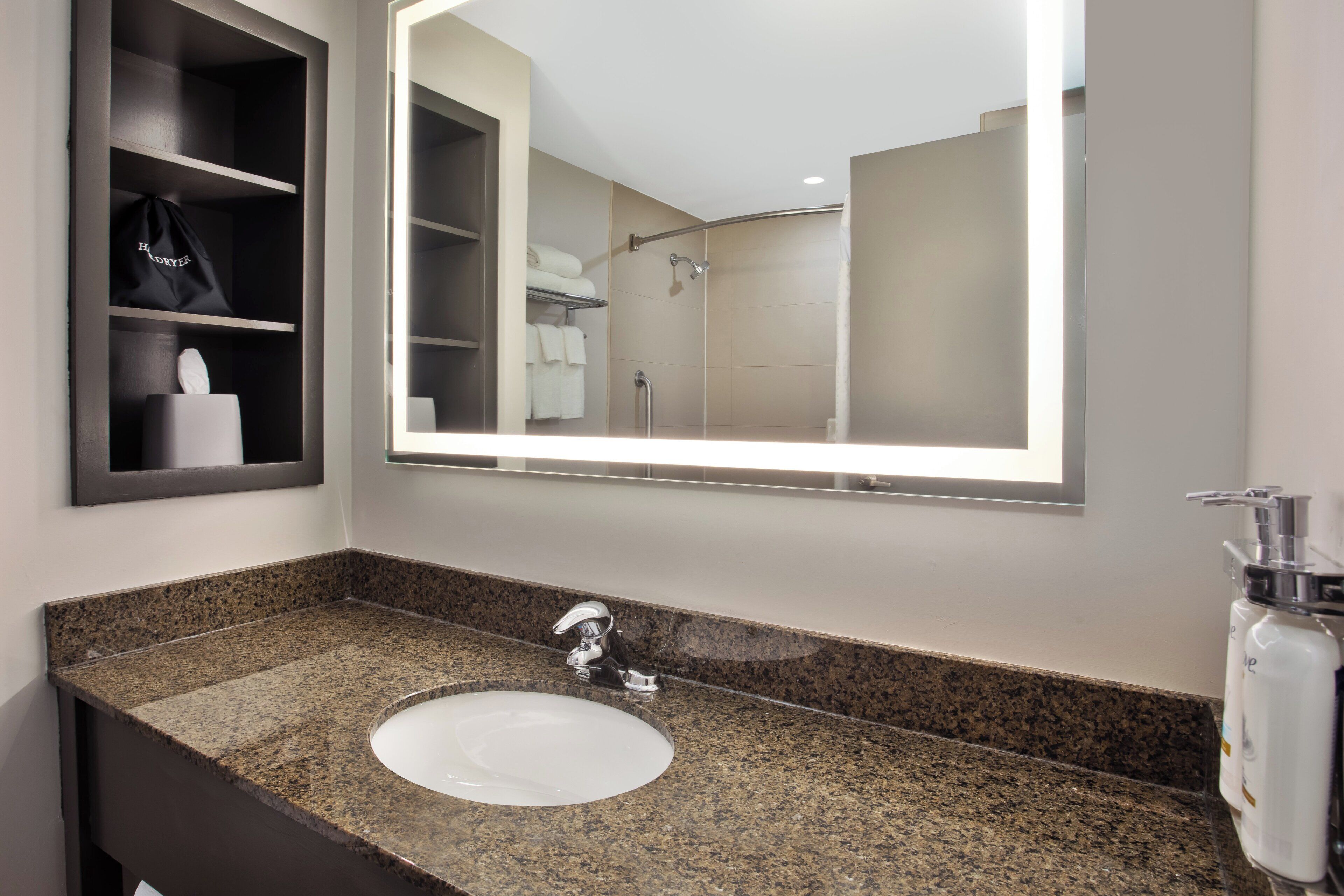 A bathroom with a granite countertop sink, a mirror, a towel rack, and a shelf with a bag and a bottle.