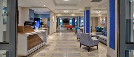 A modern hotel lobby with a reception desk, seating area, and a large TV screen.