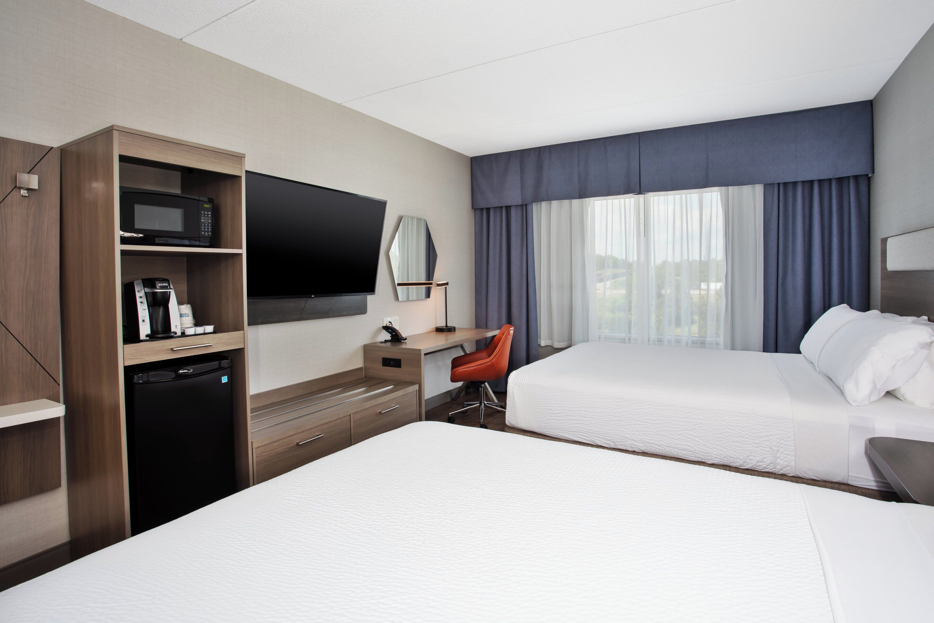 A hotel room with two beds, a desk with a flat-screen TV, a microwave, a coffee maker, and a red chair.