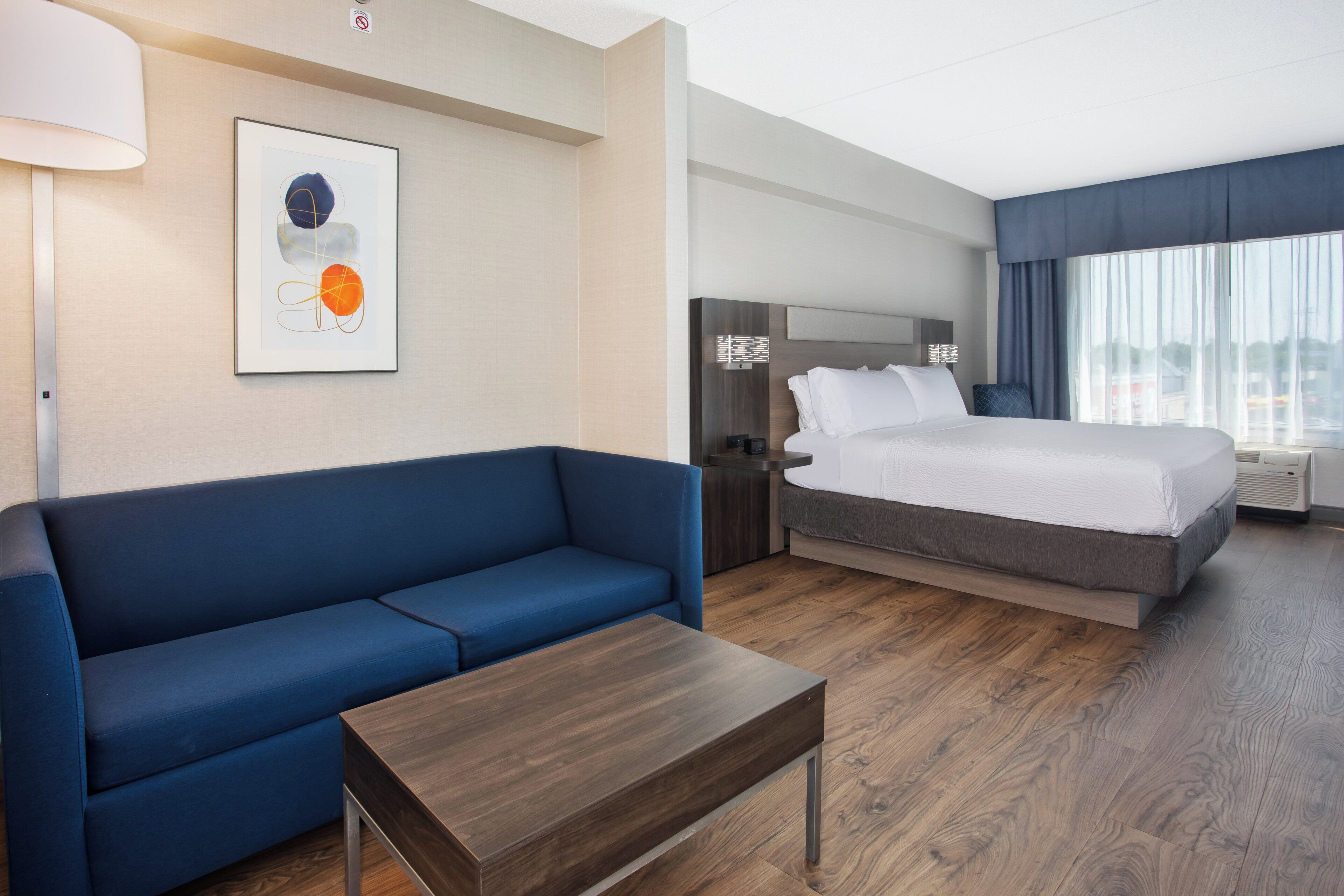Ein Hotelzimmer mit blauer Couch, einem Holztisch, einem Bett mit weißen Bettwäsche, einem Nachttisch mit Lampe und einem großen Fenster mit Vorhängen.