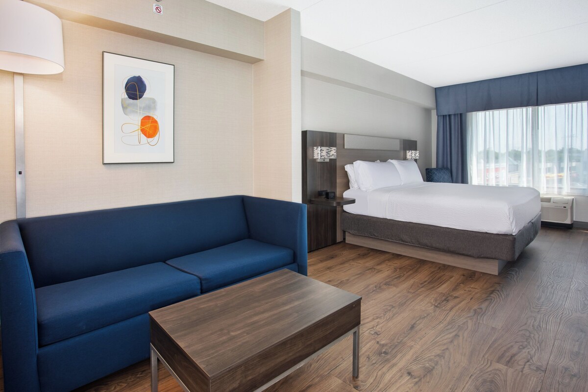 A hotel room with a blue sofa, a wooden coffee table, a bed with white bedding, a nightstand, and a wall-mounted artwork.