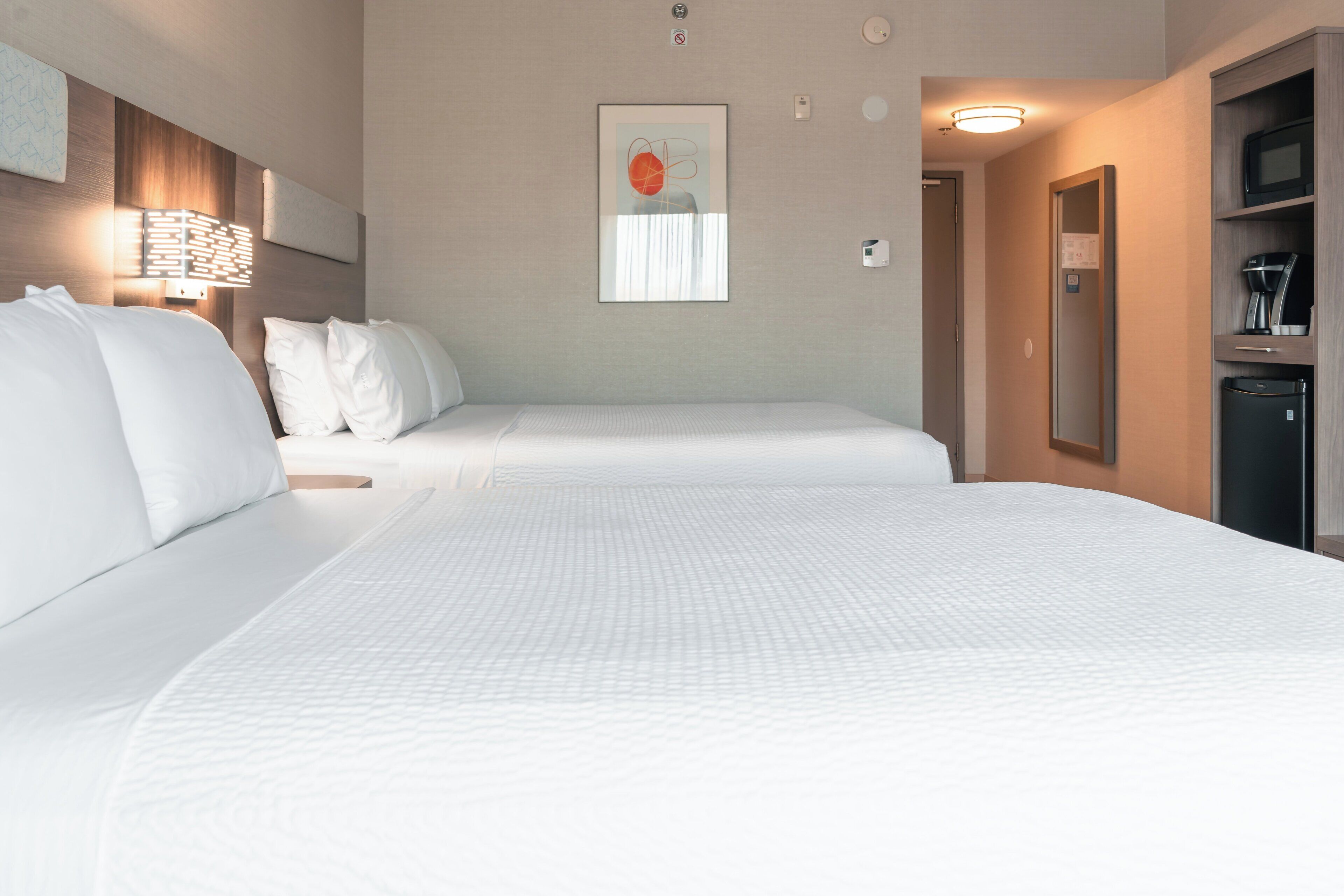 Habitación de hotel con dos camas, una obra de arte colgada en la pared, una mesita de noche, un espejo y una pequeña kitchenette.