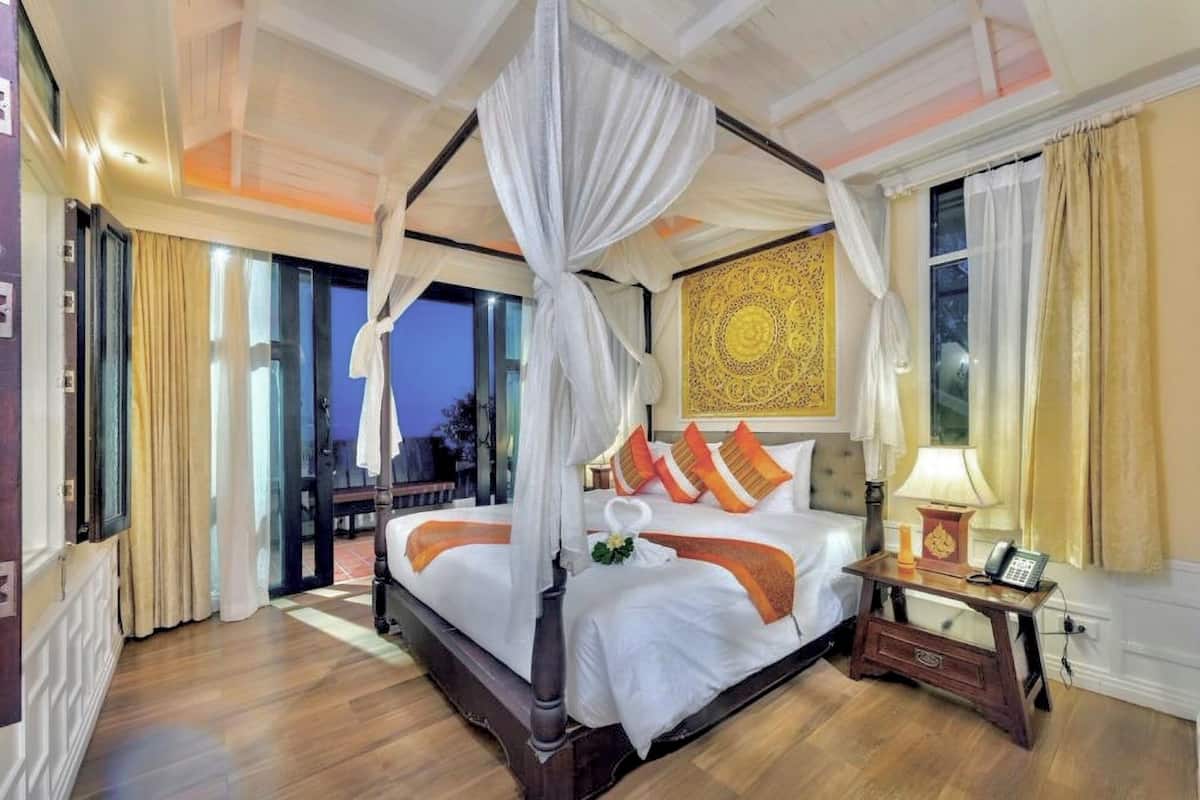 A four-poster canopy bed with white linens and orange accents, a bedside table with a lamp, and a view of the outdoors.