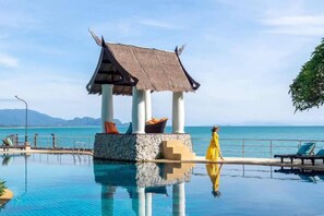 A pool with a thatched gazebo, lounge chairs, and a view of the sea.