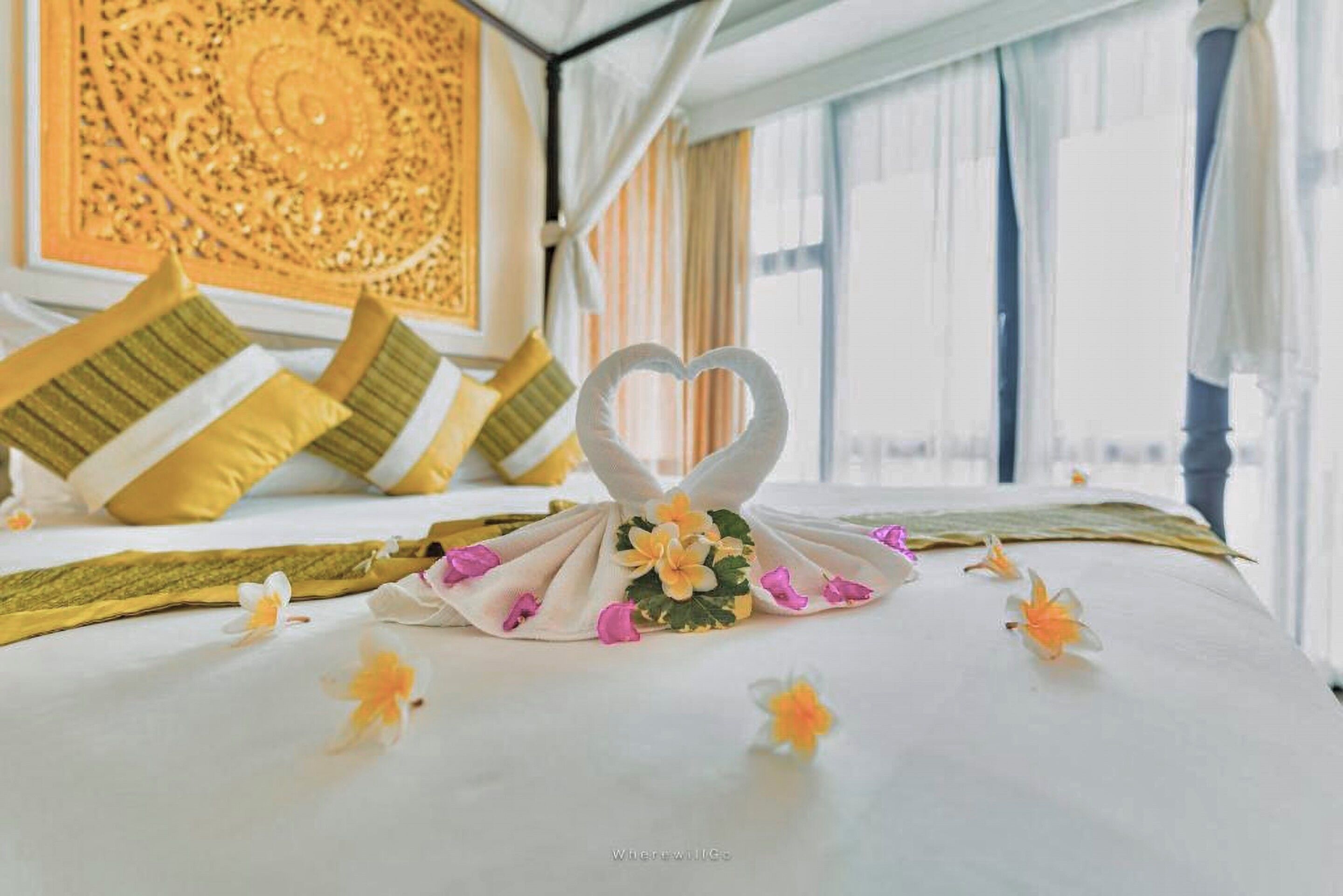 A neatly made bed with a heart-shaped towel and flowers, yellow pillows, and a decorative wall piece.