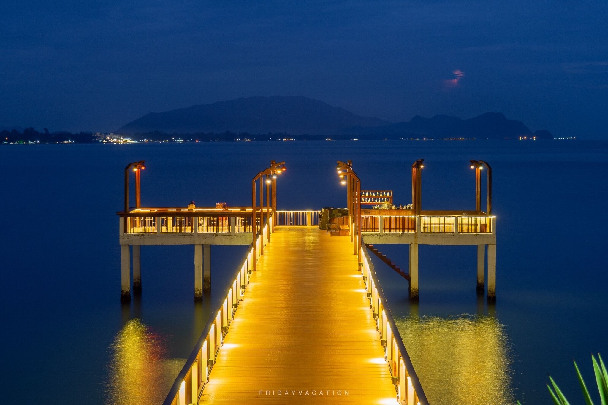 A long, illuminated pier extending into calm waters at night, with a mountain in the background.