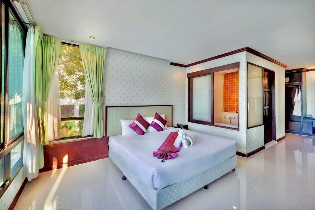 A hotel room with a large bed, a bathroom with a glass shower, and a view of greenery outside.