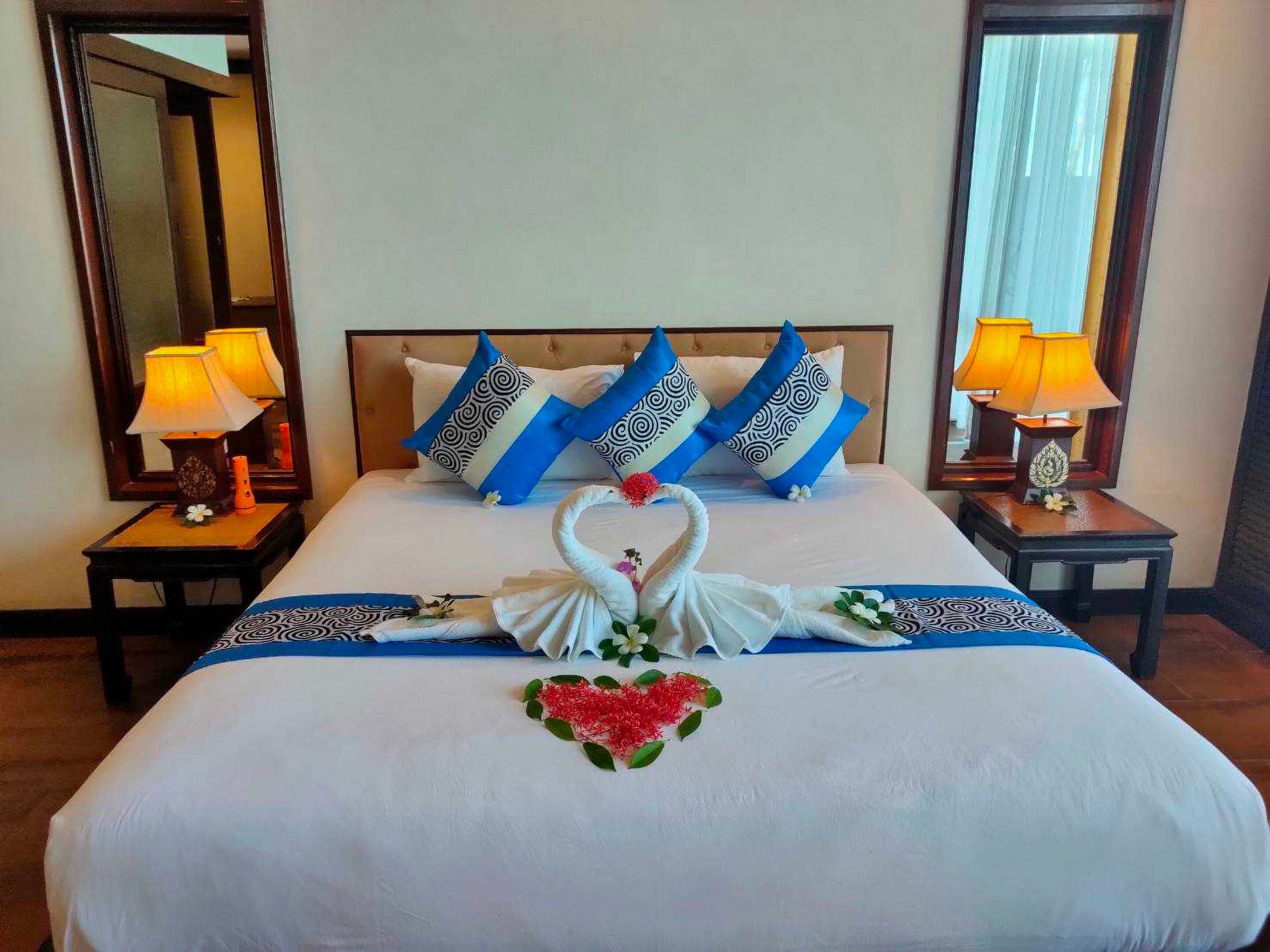 Una cama bien hecha con decoración de cisne y corazón, almohadillas estampadas en azul y blanco y dos lámparas de noche.
