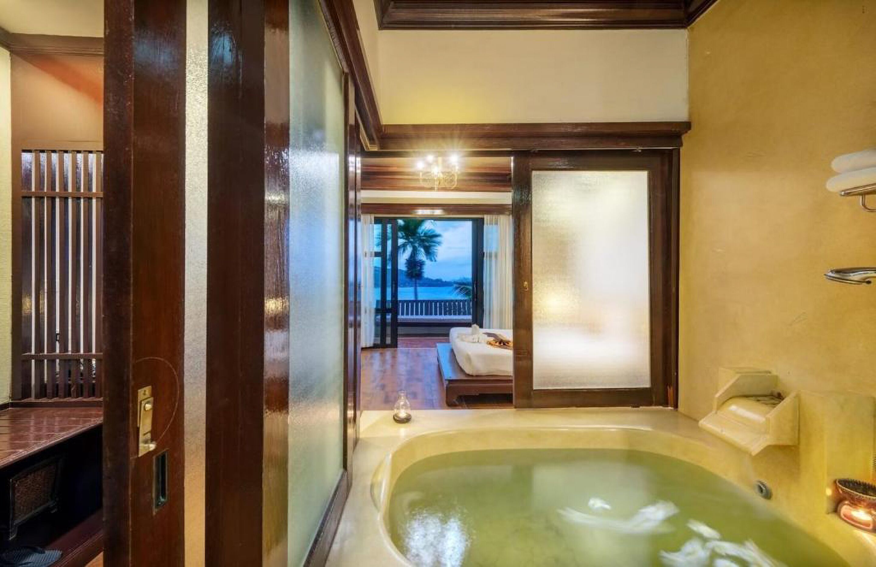 Ein luxuriöses Badezimmer mit einer großzügigen Badewanne, einer Holztür und Blick auf einen Balkon mit einer Palme.