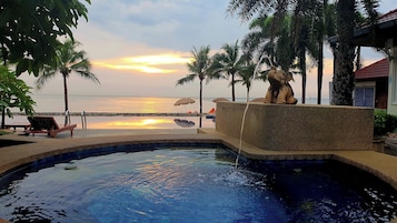 A pool with a stone statue, palm trees, and a view of the sunset over the water.