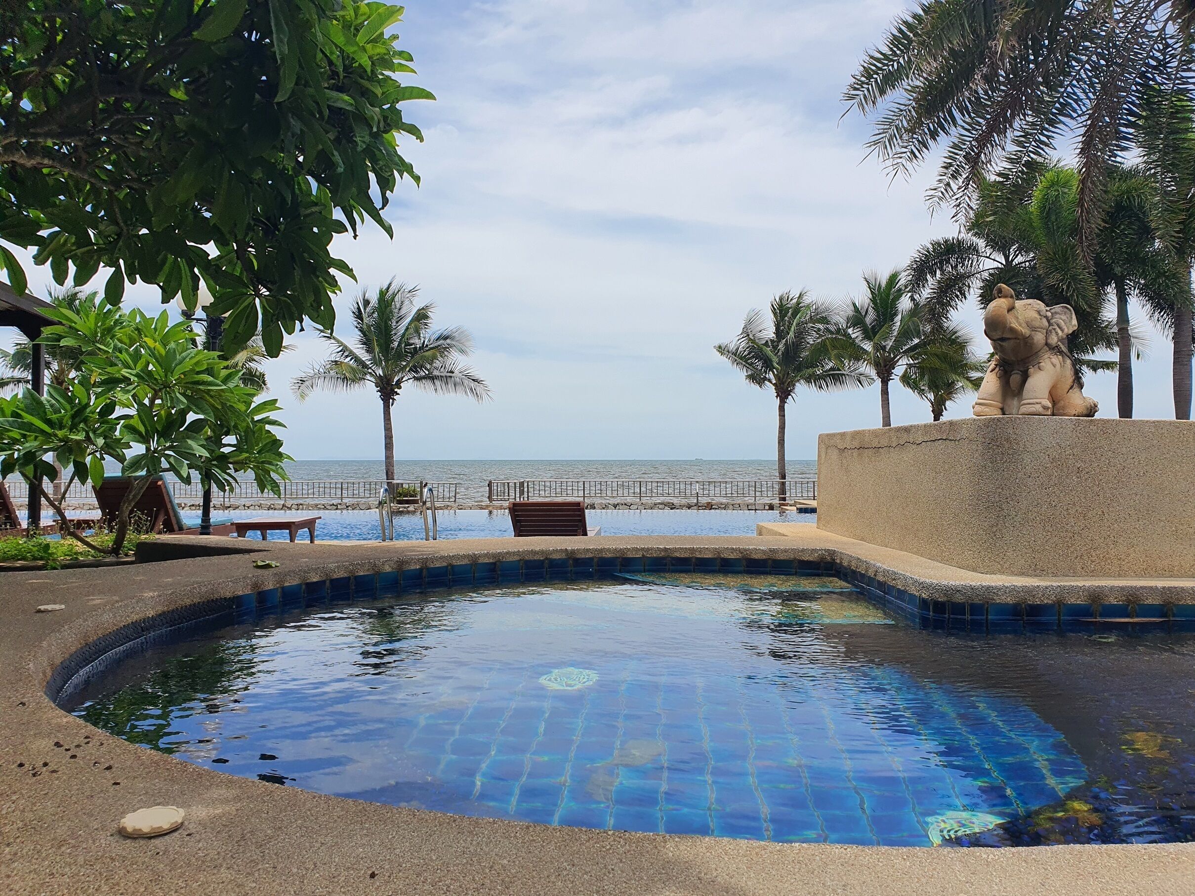 A swimming pool with a view of the sea, surrounded by palm trees and a statue.
