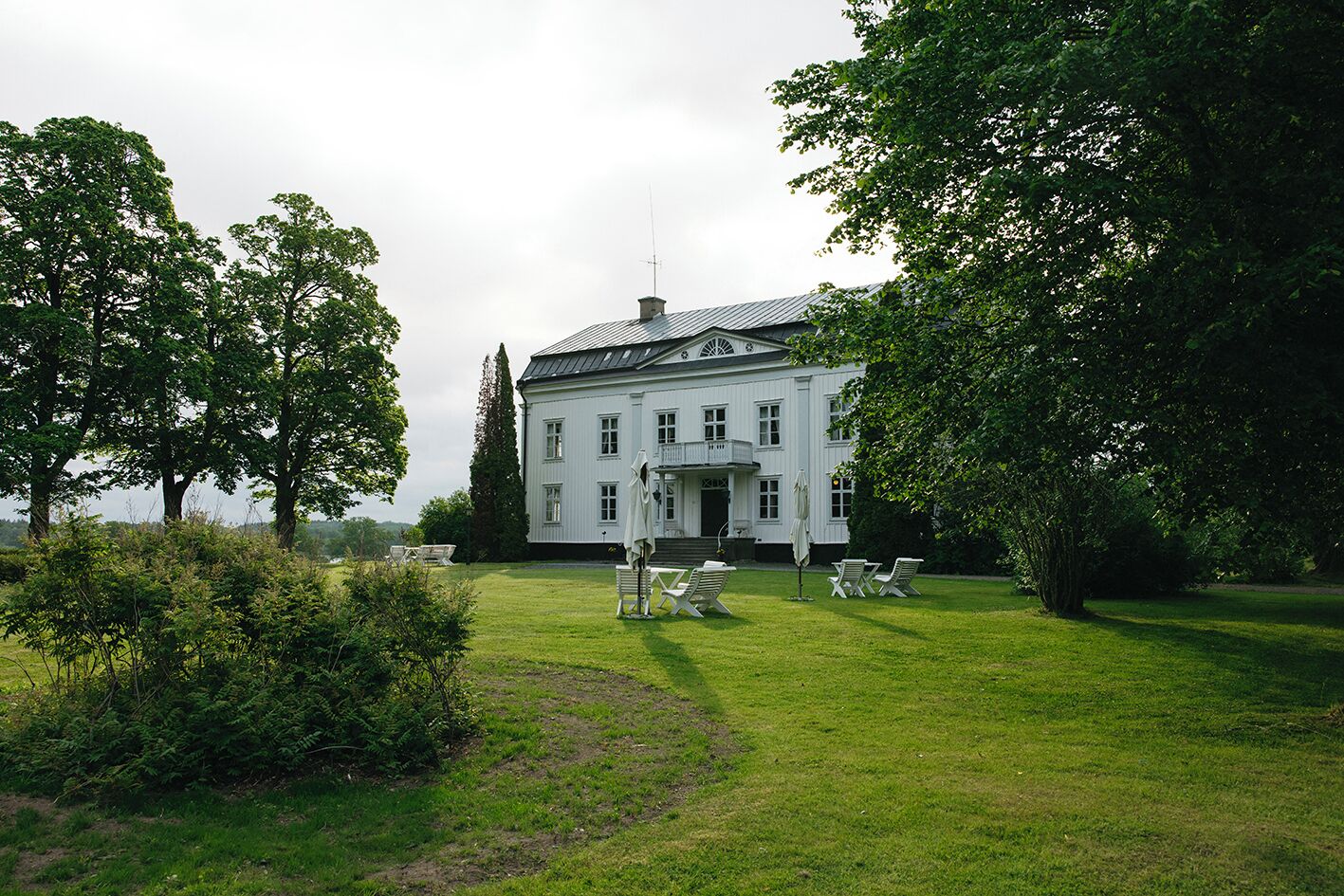 A large white building with a symmetrical facade, a central entrance with steps, and a surrounding lawn with chairs and umbrellas.