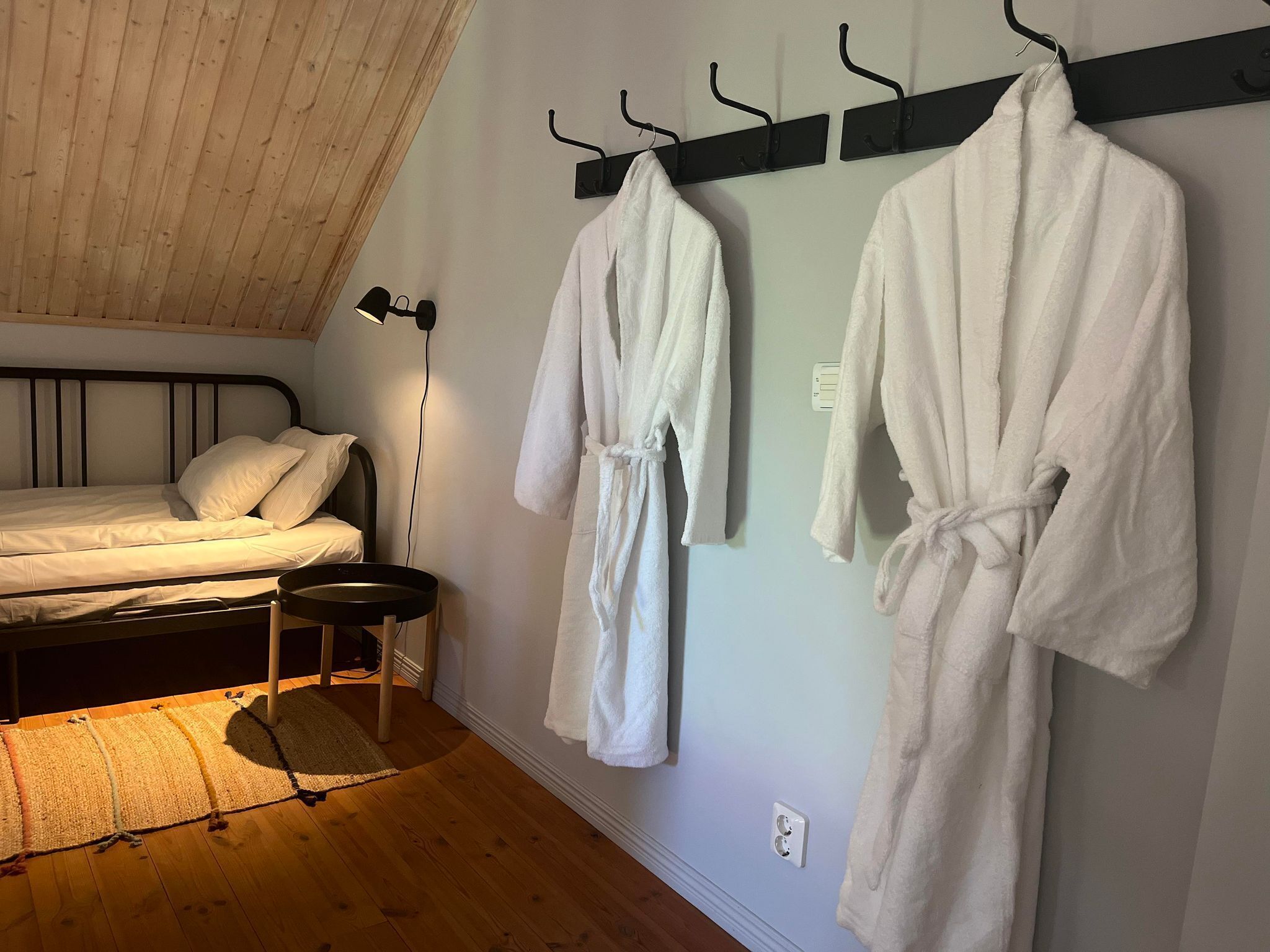 A bedroom with a bed, a small bedside table, a lamp, and two white bathrobes hanging on hooks.