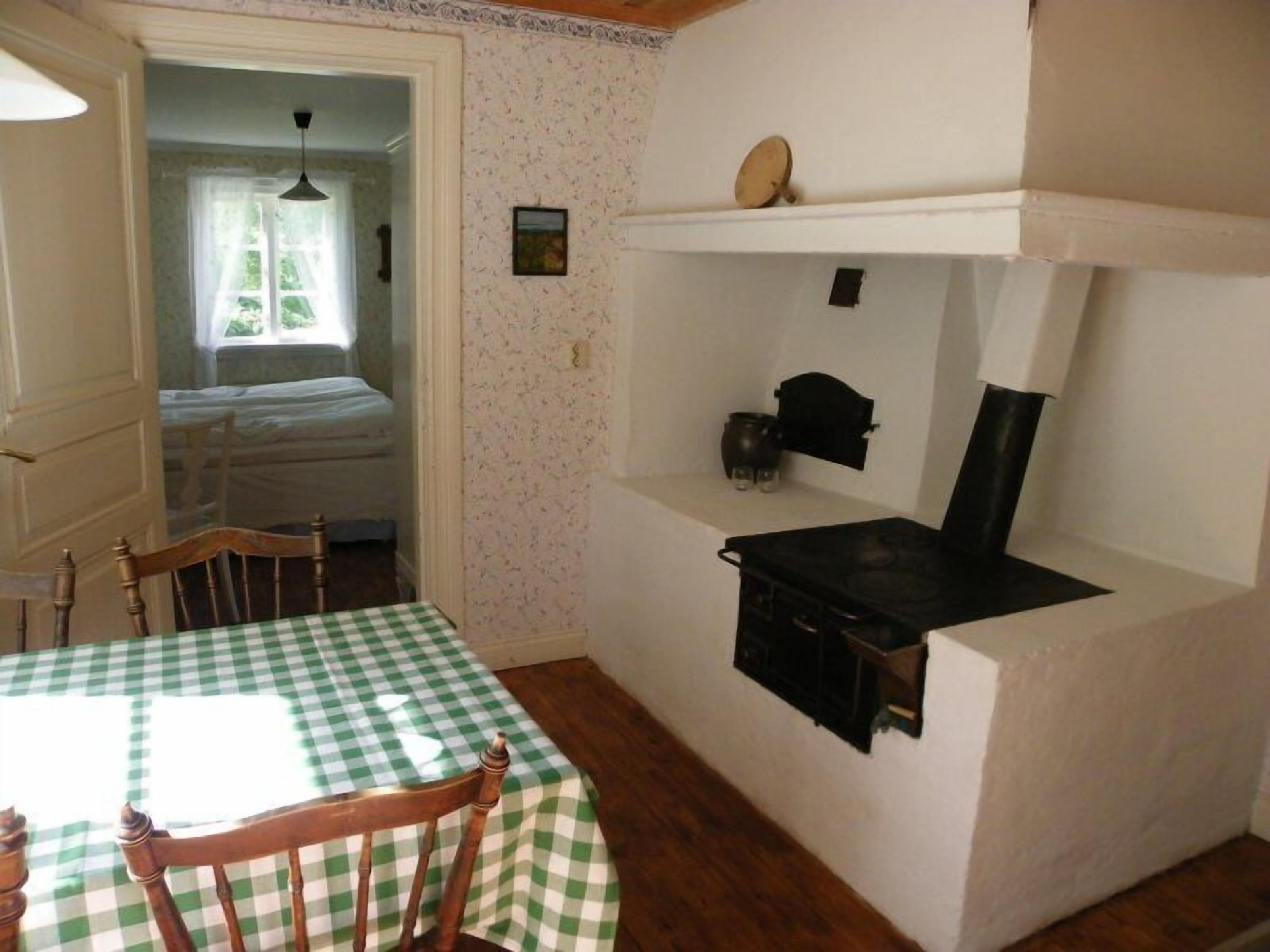 A room with a checkered table, a bed, a shelf with a kettle, and a wall clock.