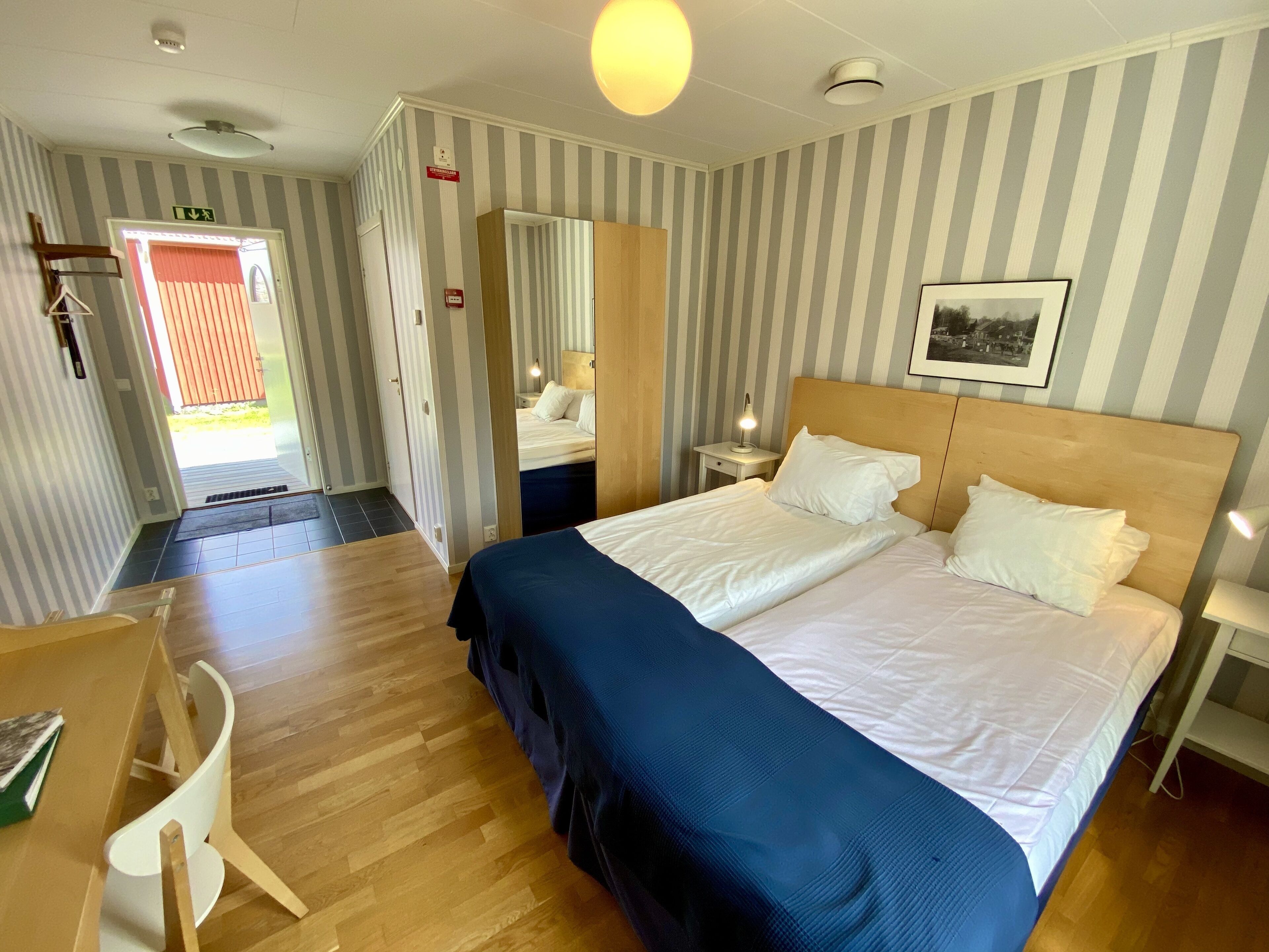 A hotel room with a double bed, a desk with a chair, a bedside table with a lamp, and a door leading to a balcony.