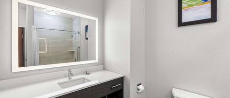 Un baño moderno con un espejo grande, lavamanos blanco, tocador oscuro y un inodoro.
