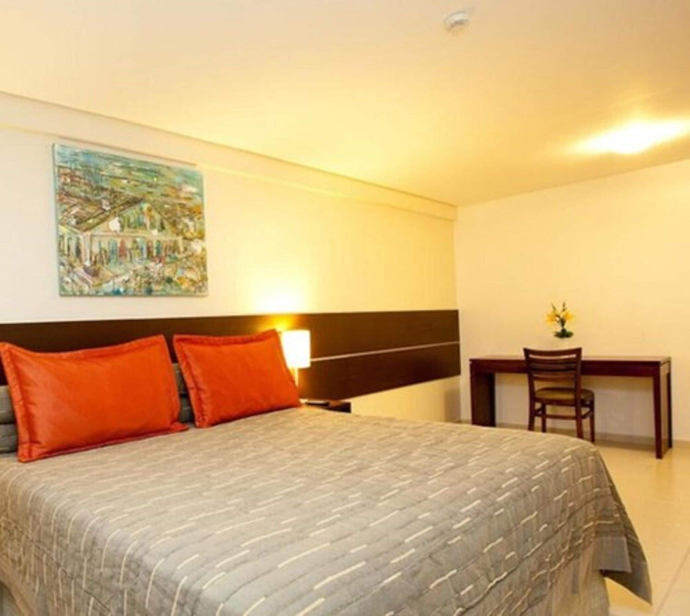 A hotel room with a bed, two orange pillows, a nightstand, a desk with a chair, and a painting on the wall.